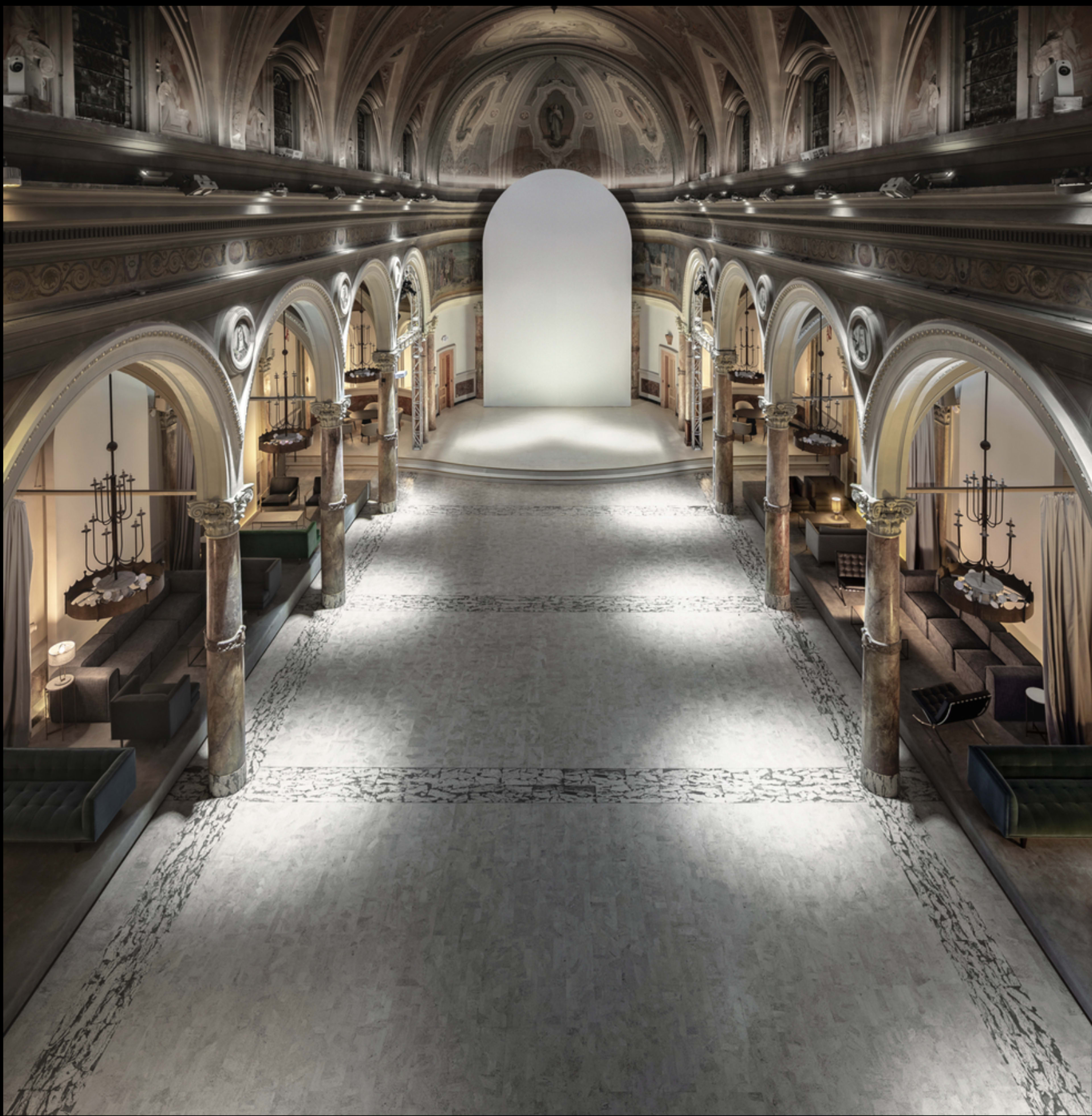 A spacious interior hall with tall arches, decorative columns, and a minimalistic stage area illuminated by soft lighting.