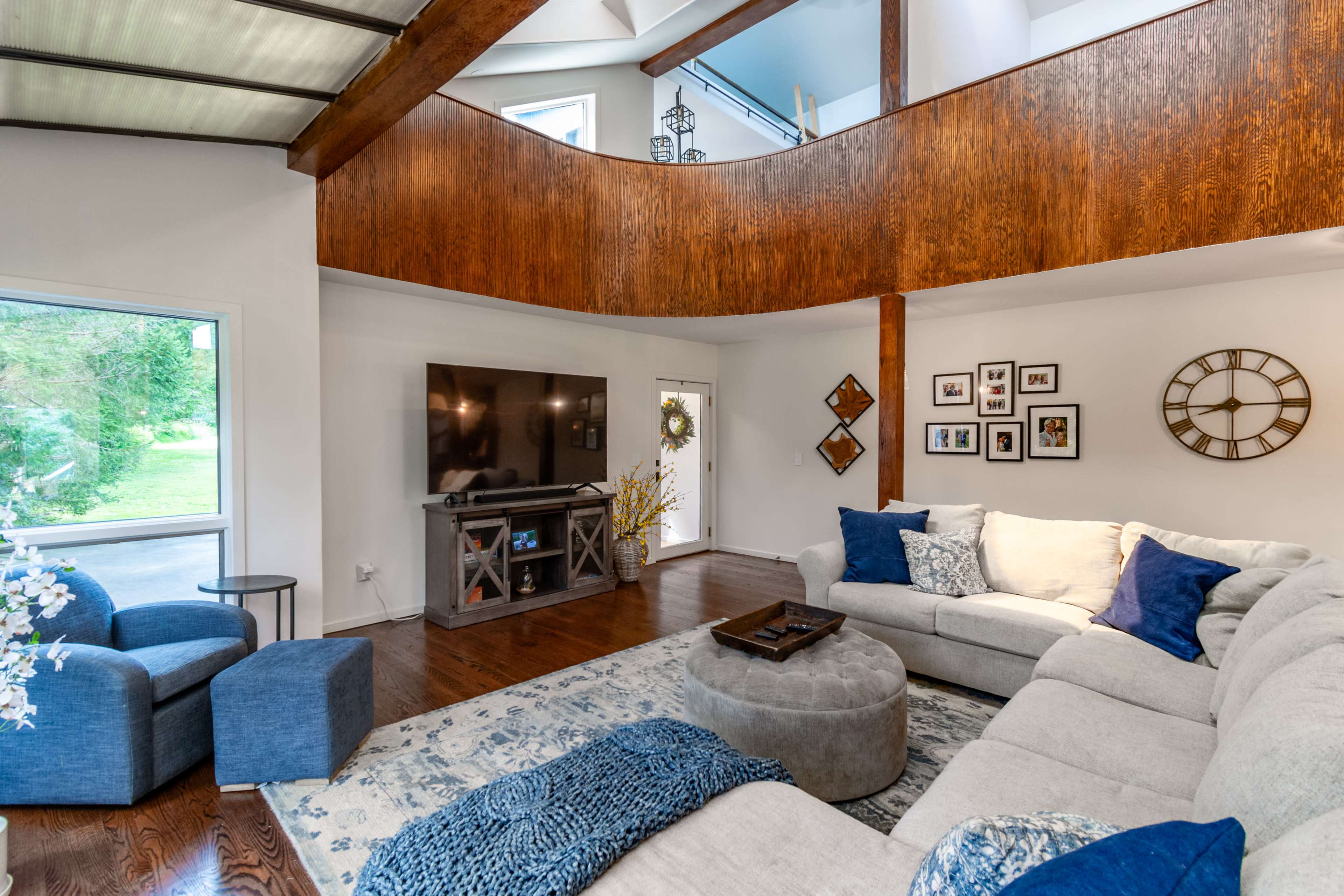 The image shows a spacious living room featuring a large sectional sofa, a coffee table, and a TV stand, with large windows providing natural light and a view of the outdoors.