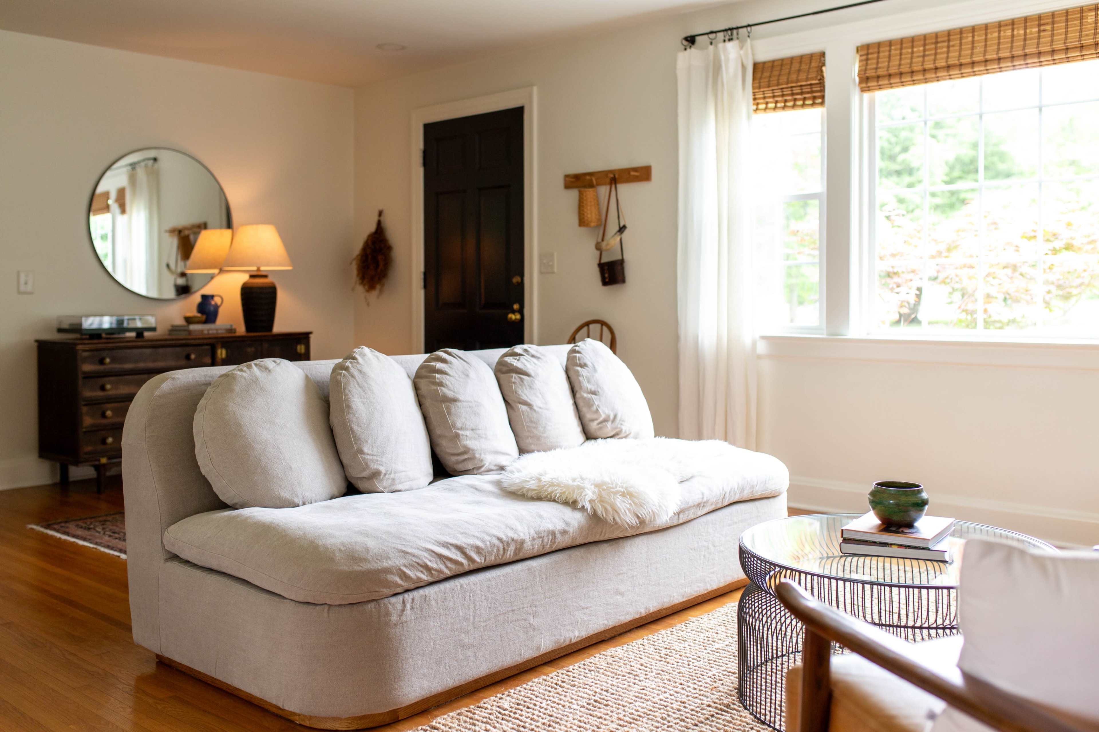 The living room features a gray sofa with five cushions, a round mirror, a wooden dresser, and a large window with curtains.