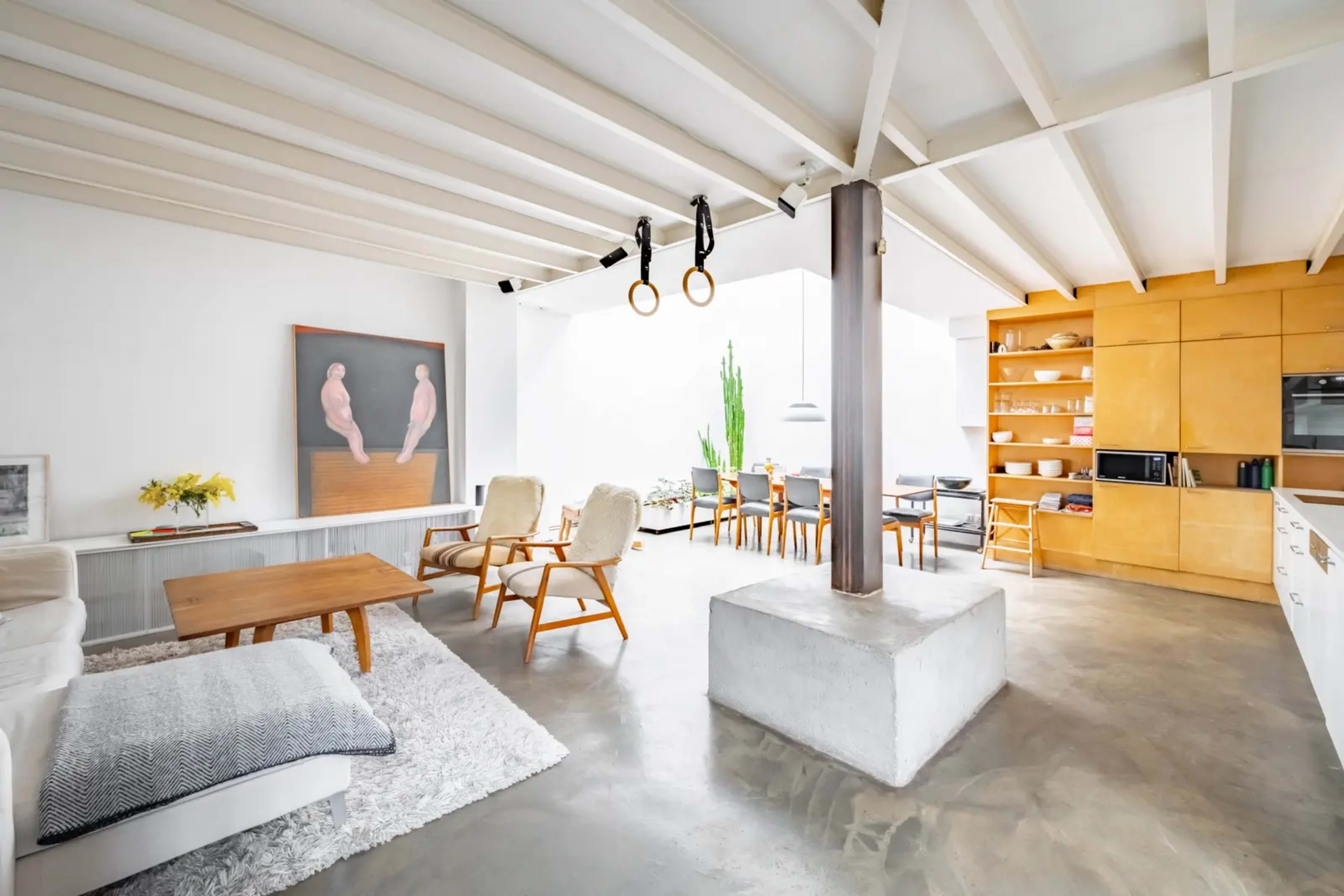 The image depicts a modern, open-concept living area featuring a dining space, a seating area with chairs, and a large piece of artwork on the wall.