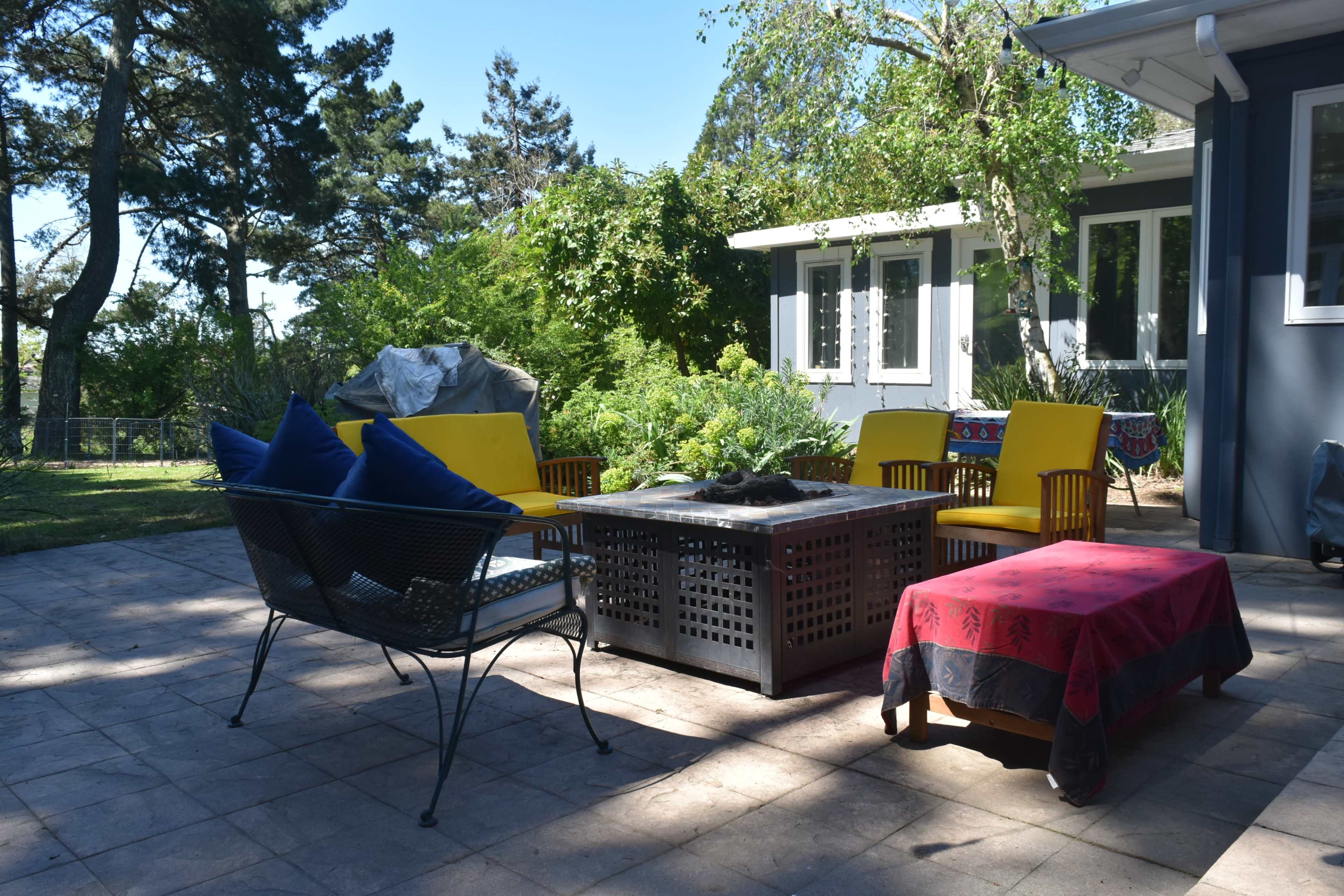 A patio features a table with a fire pit surrounded by yellow chairs and a black wire sofa, with greenery and a house in the background.