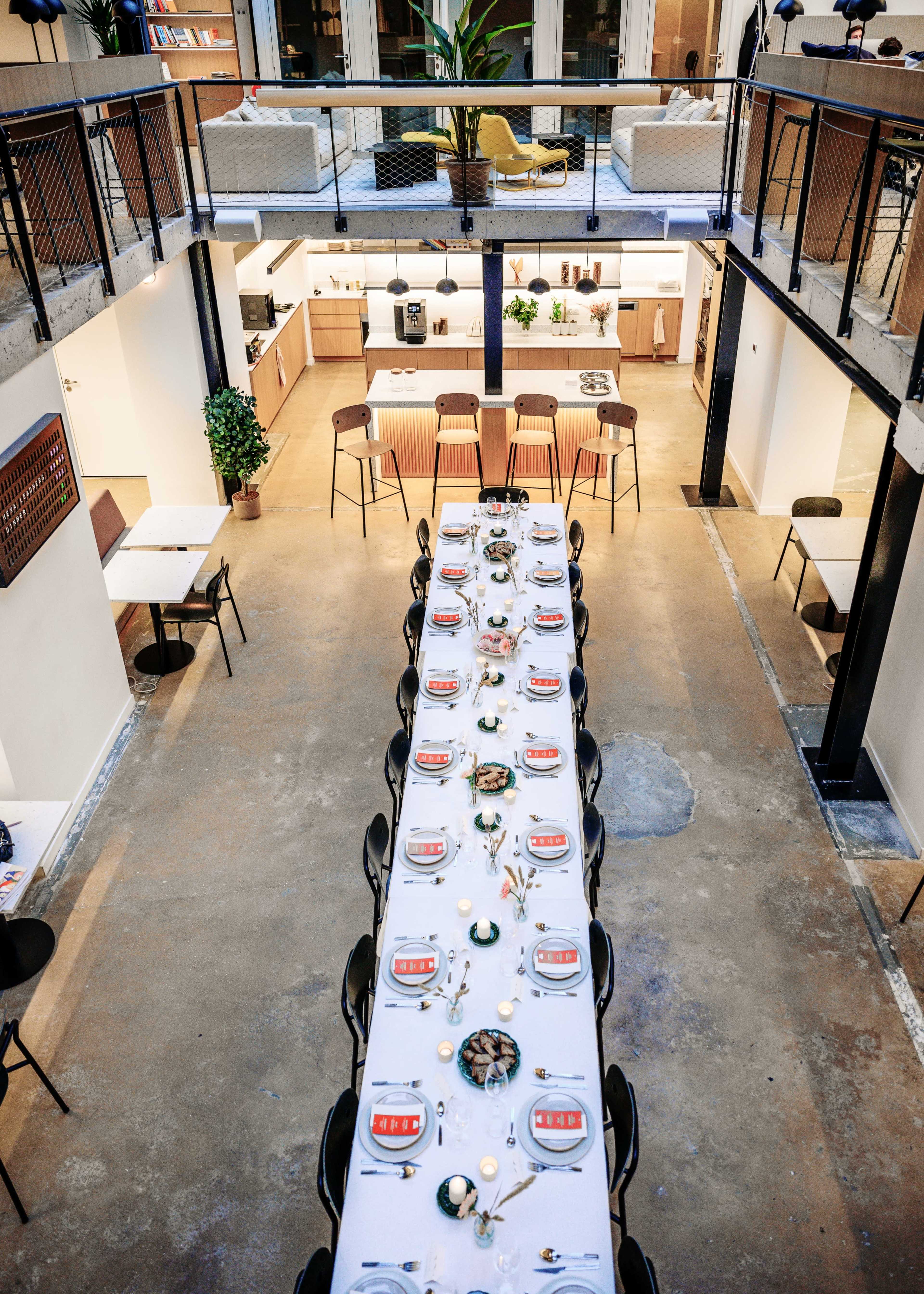 A long, neatly set dining table is positioned in a spacious, modern kitchen area with a balcony overlooking the seating arrangement.