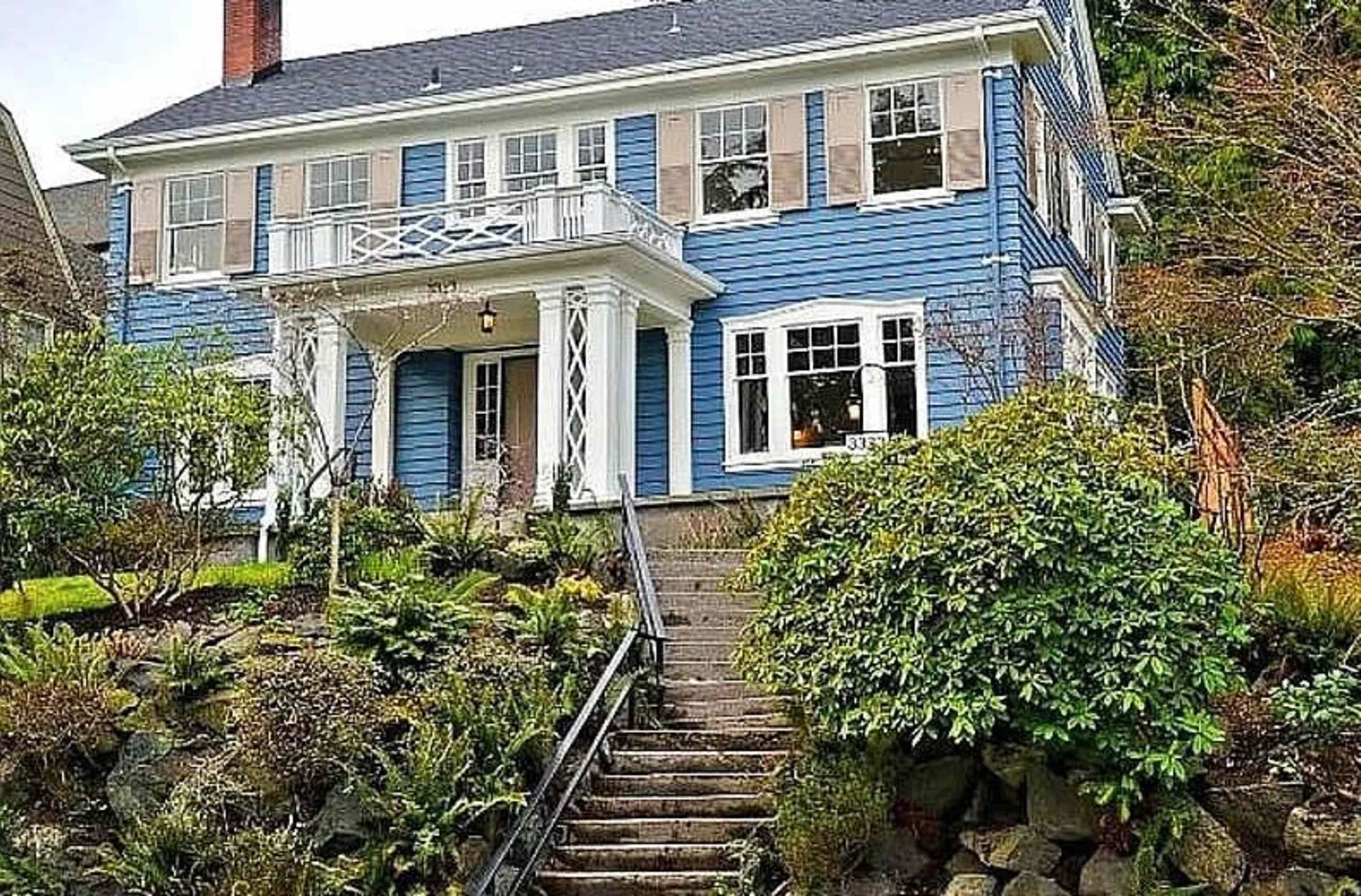 A two-story blue house with white trim, featuring a front porch and a staircase leading up to it, surrounded by greenery.