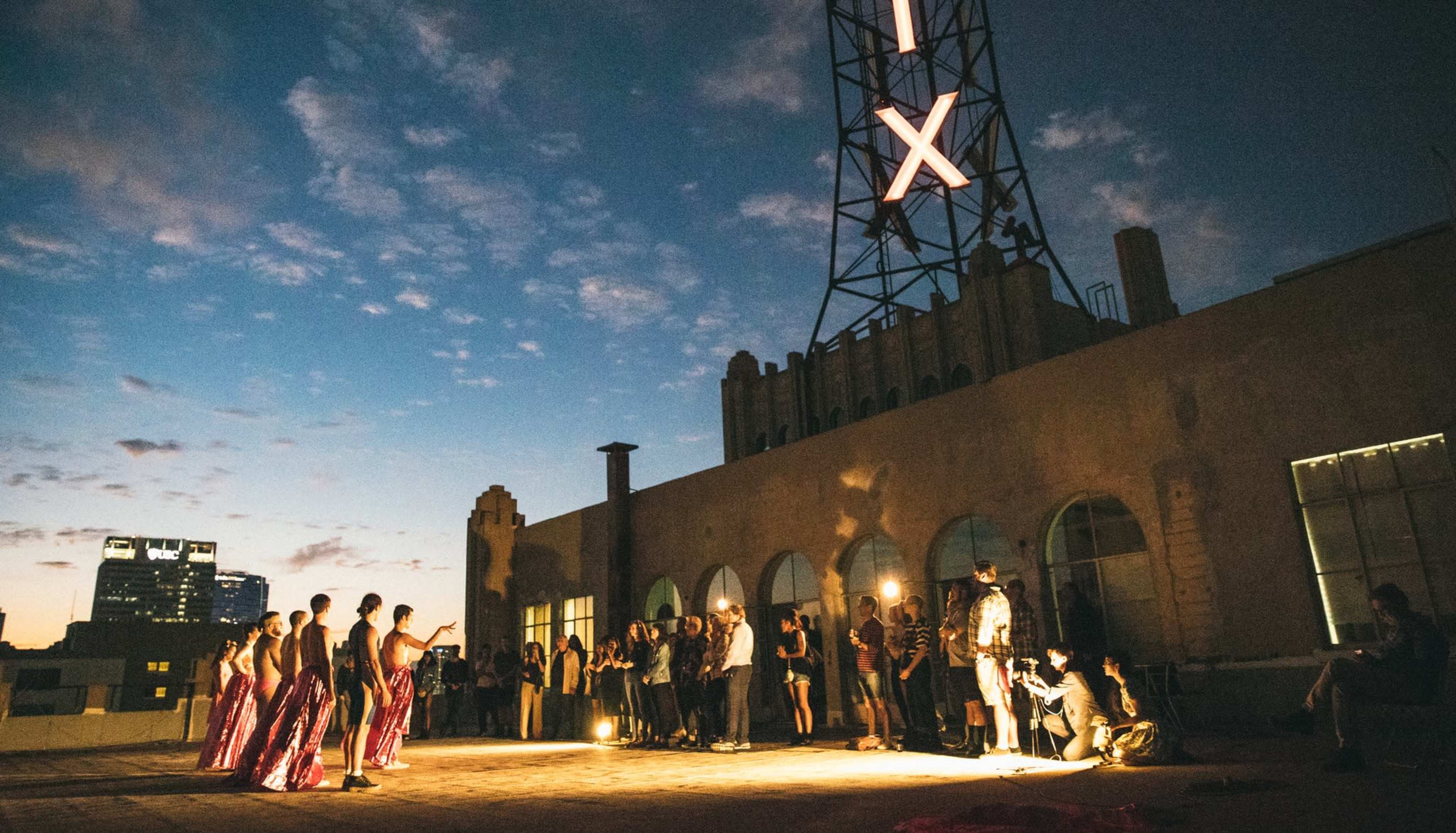 Models in colorful outfits pose on a rooftop during a photoshoot as a crowd of photographers and onlookers watch in the fading light.