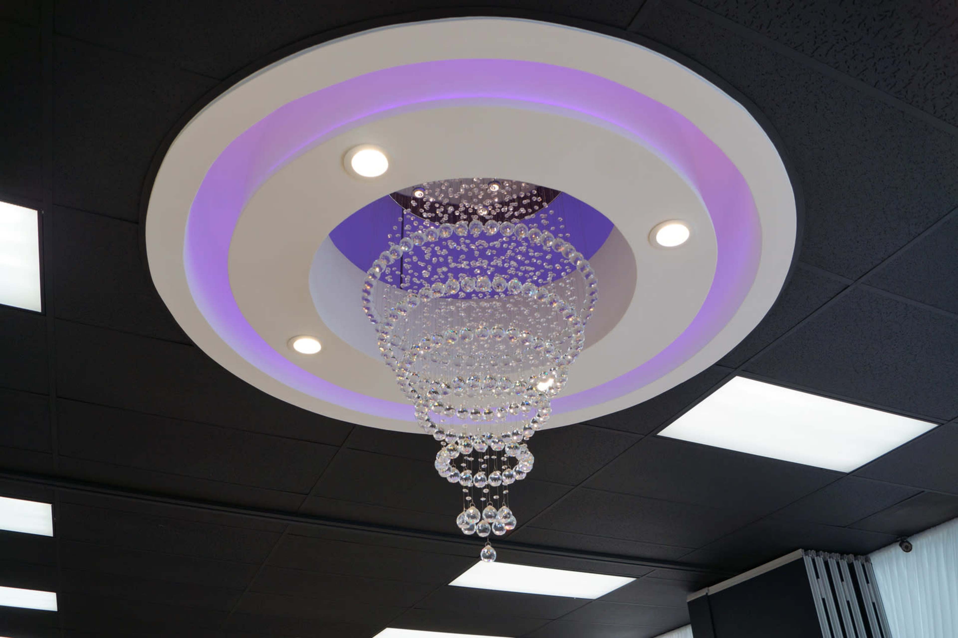 The image shows a modern ceiling light fixture with a series of chandeliers hanging from a circular ceiling design, illuminated by purple and white lights.