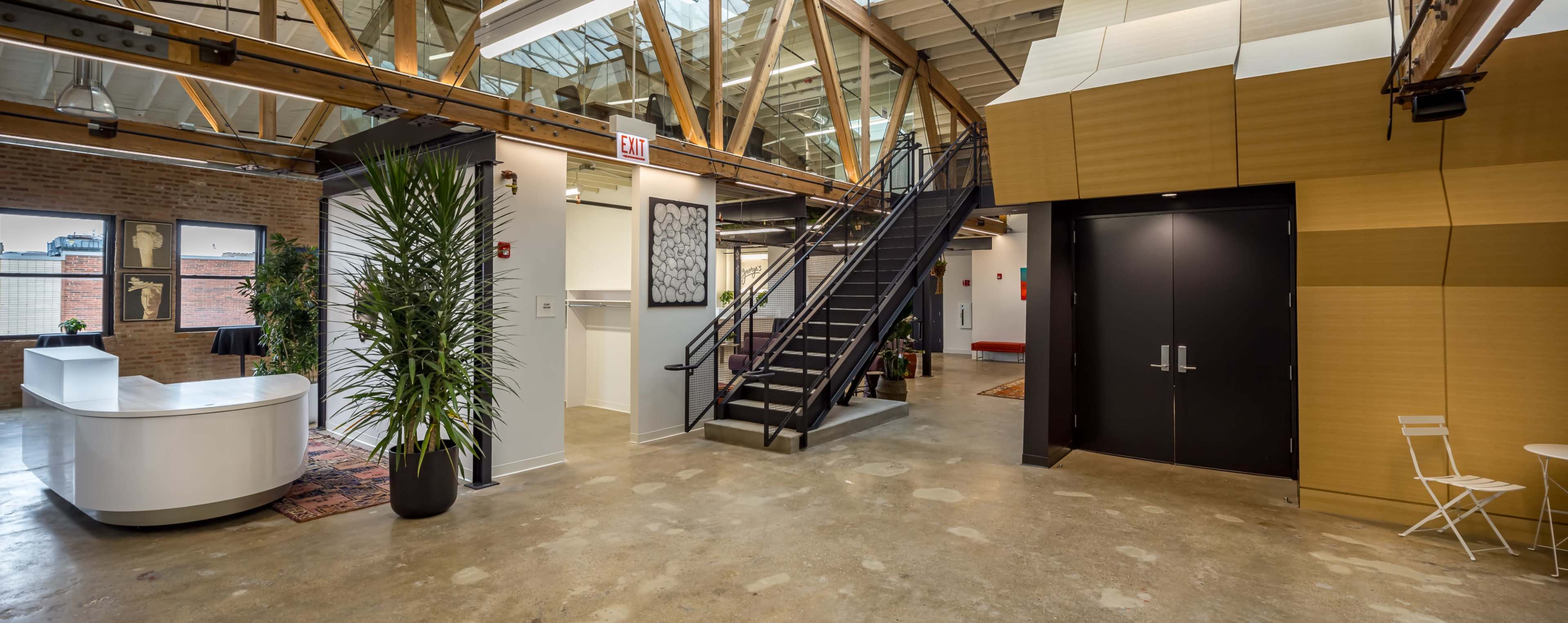 The image shows a modern office lobby featuring a reception desk, a staircase leading to an upper level, and a mix of plants and artwork on the walls.