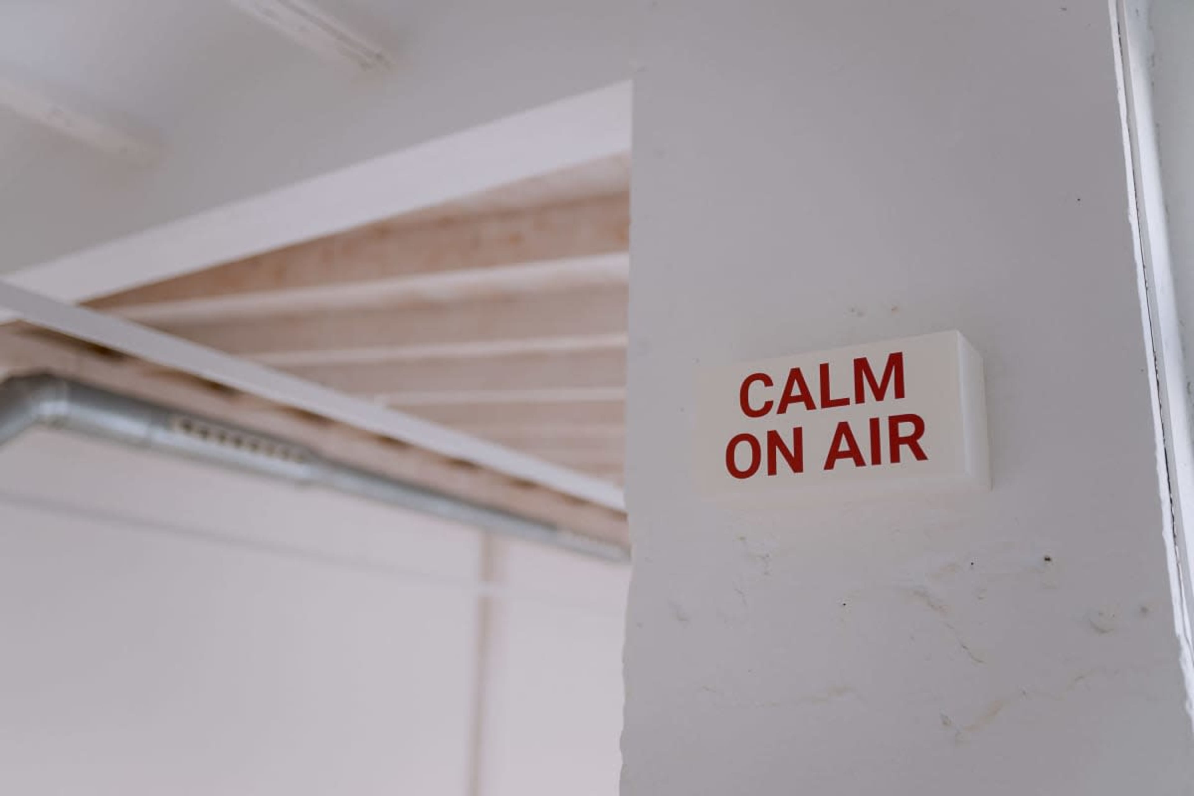 A sign reading "CALM ON AIR" is mounted on a white wall in a room with a wooden ceiling.