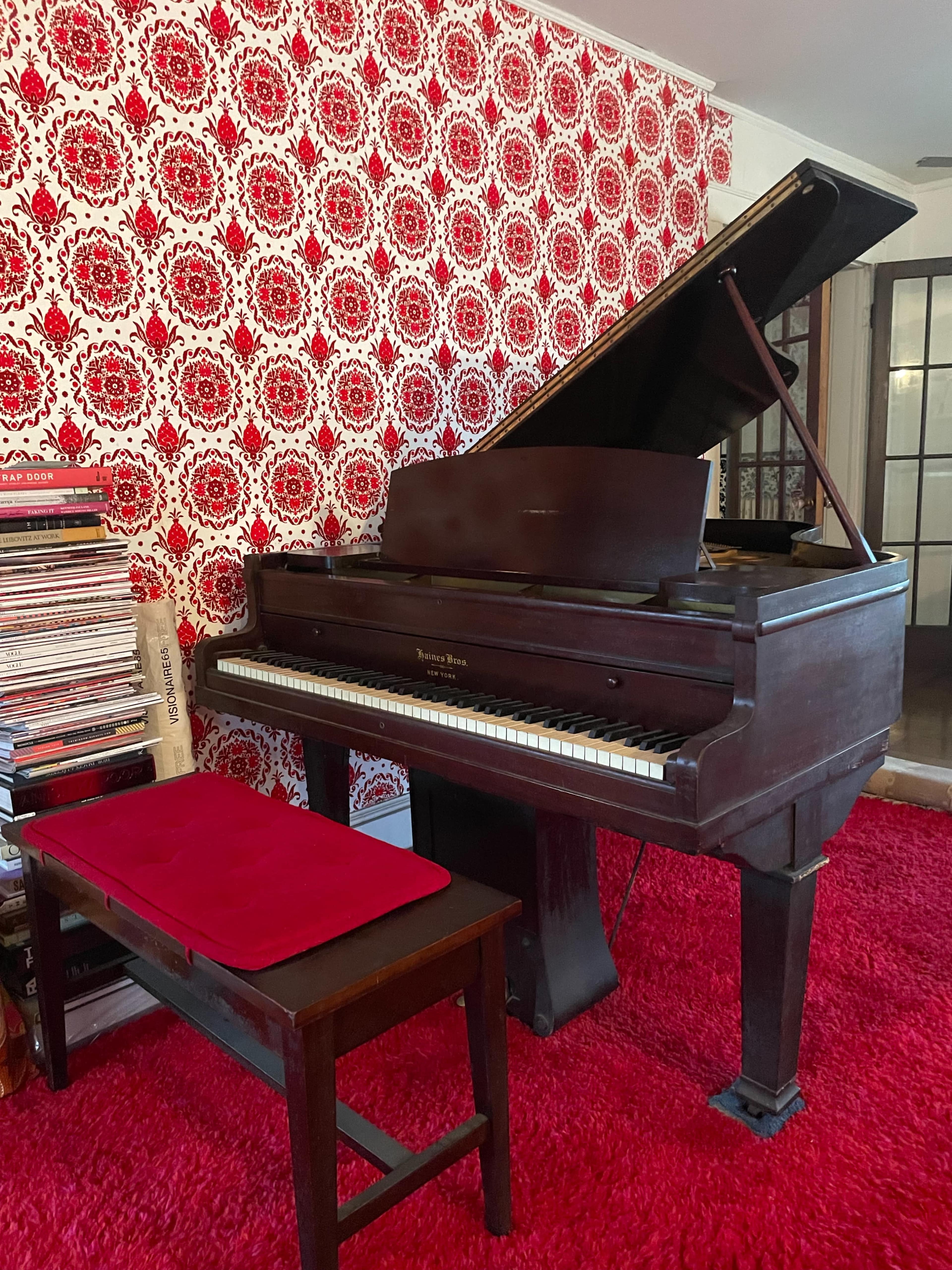 A grand piano with an open lid is positioned beside a stack of records against a wall covered in red patterned wallpaper.