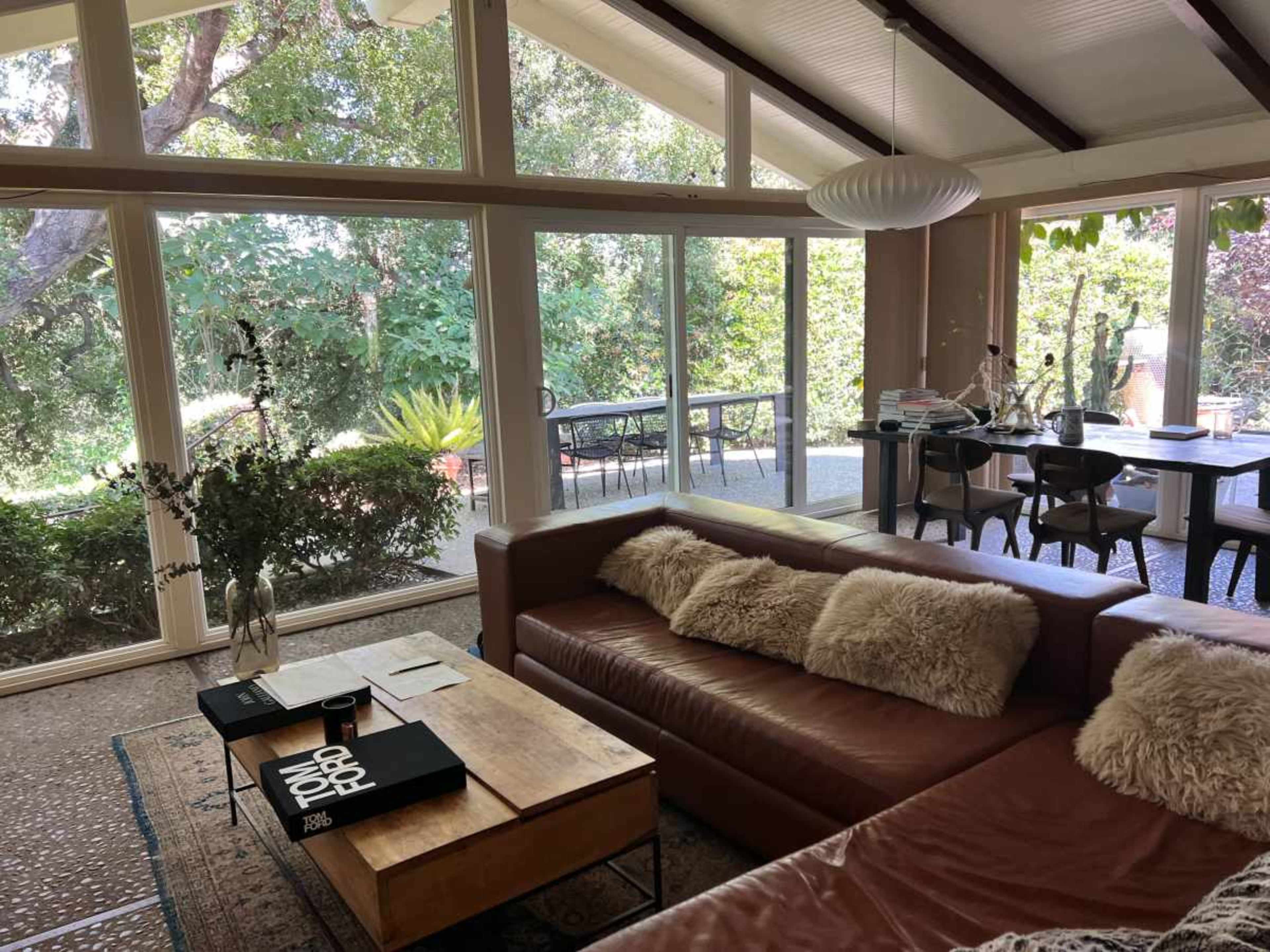 A spacious living room features a brown leather sofa, a coffee table with books, and large windows overlooking a green garden.