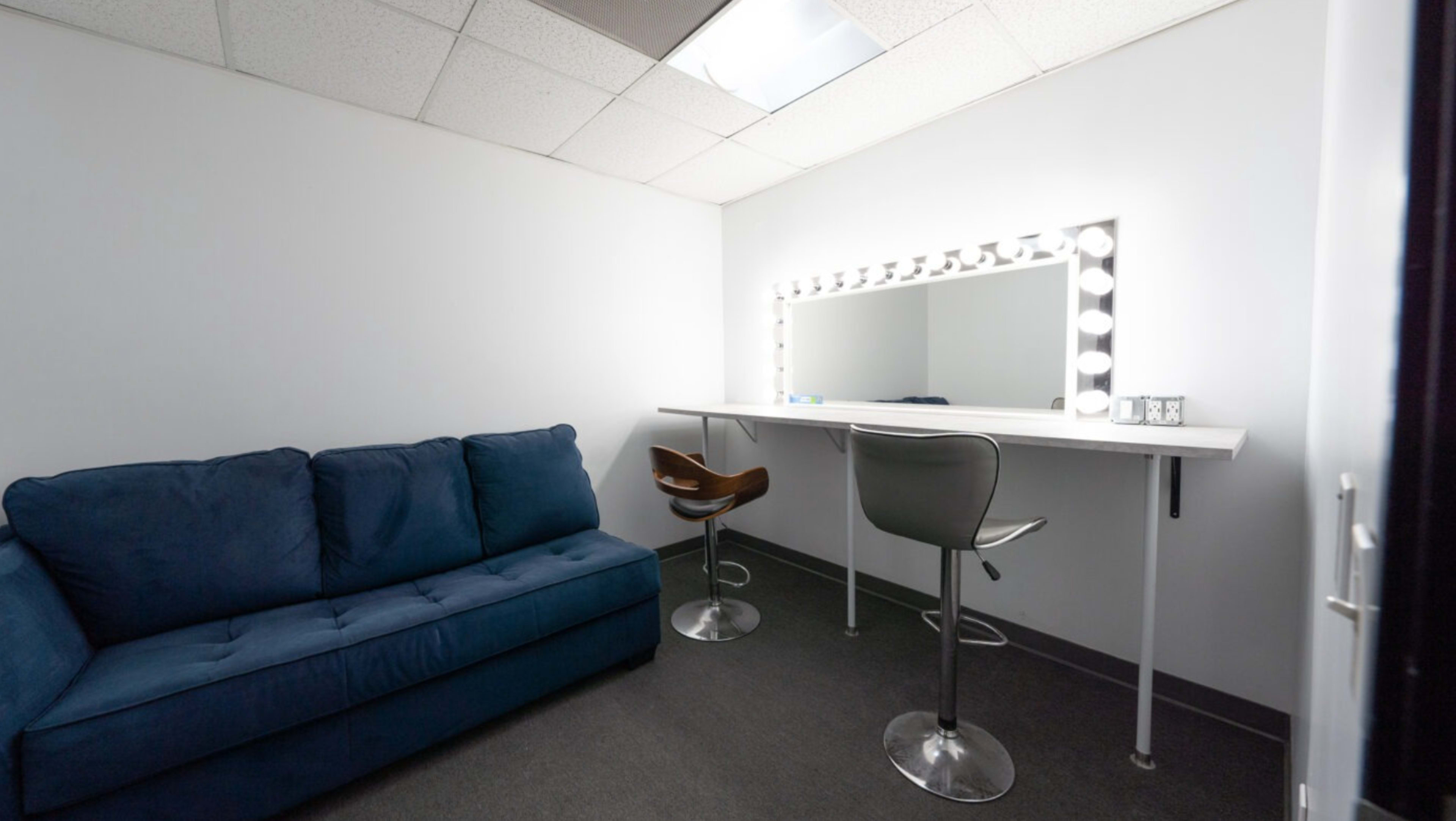 A small, well-lit room with a blue couch and a vanity area featuring a large mirror surrounded by lights.