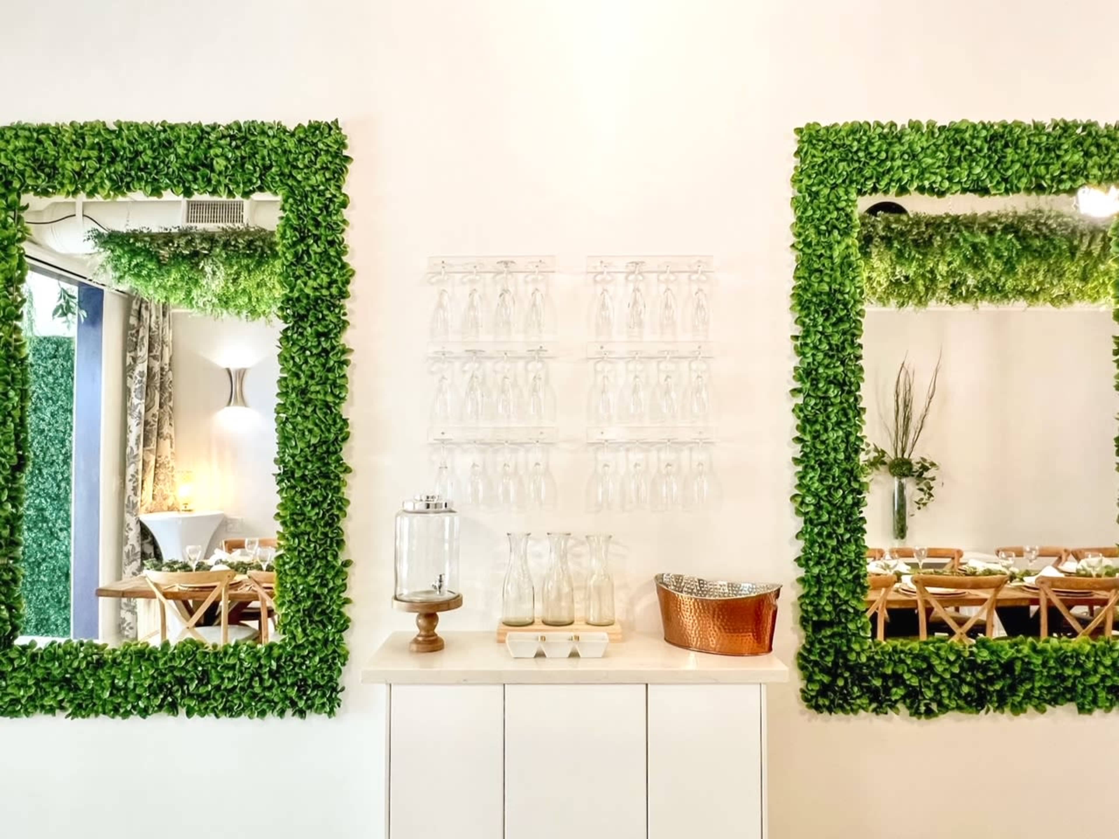 The image shows a decorated wall with two leafy green mirrors flanking a display of glass bottles on a white cabinet.
