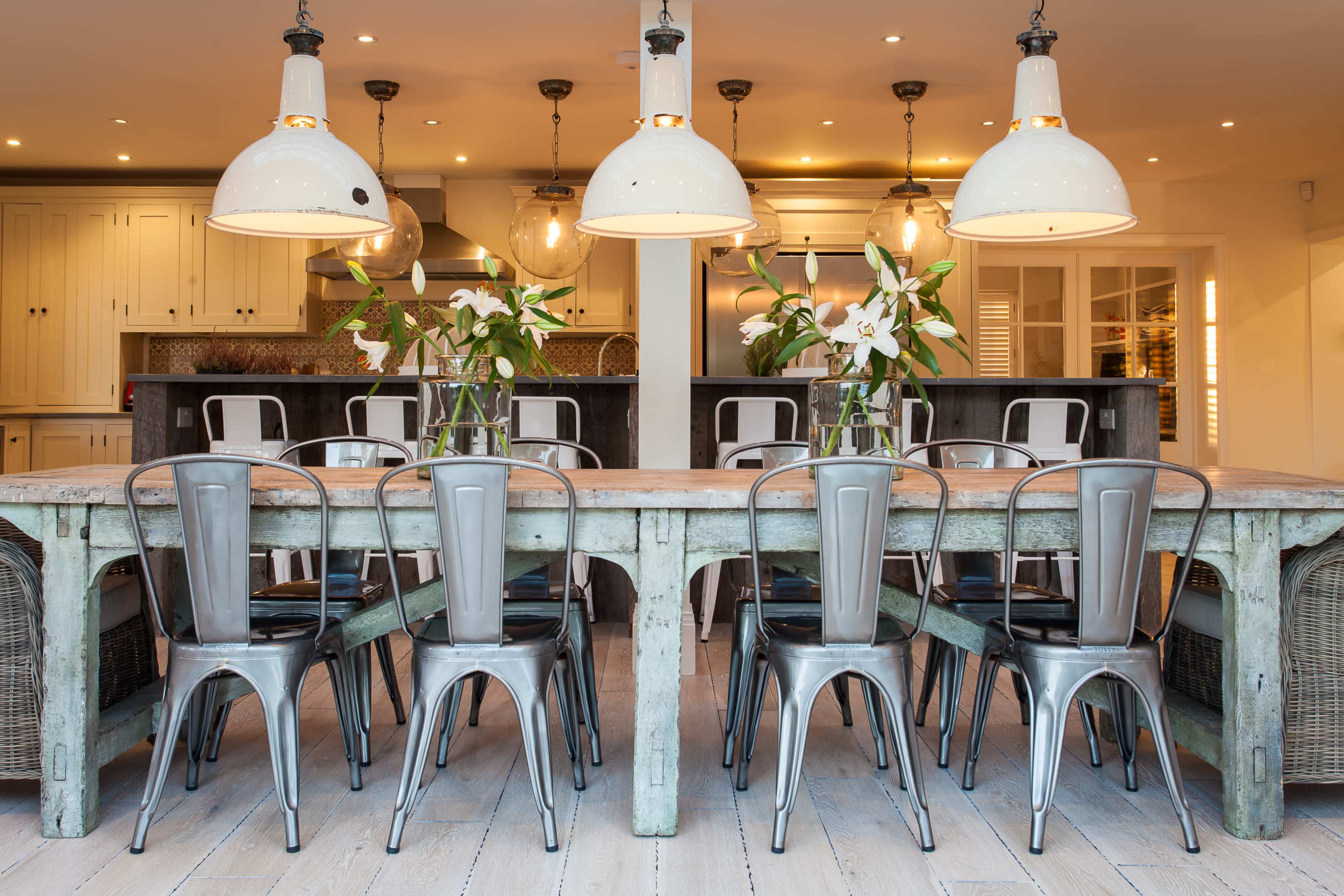 A rustic wooden dining table is surrounded by metallic chairs, with large pendant lights hanging above and floral arrangements as centerpieces.