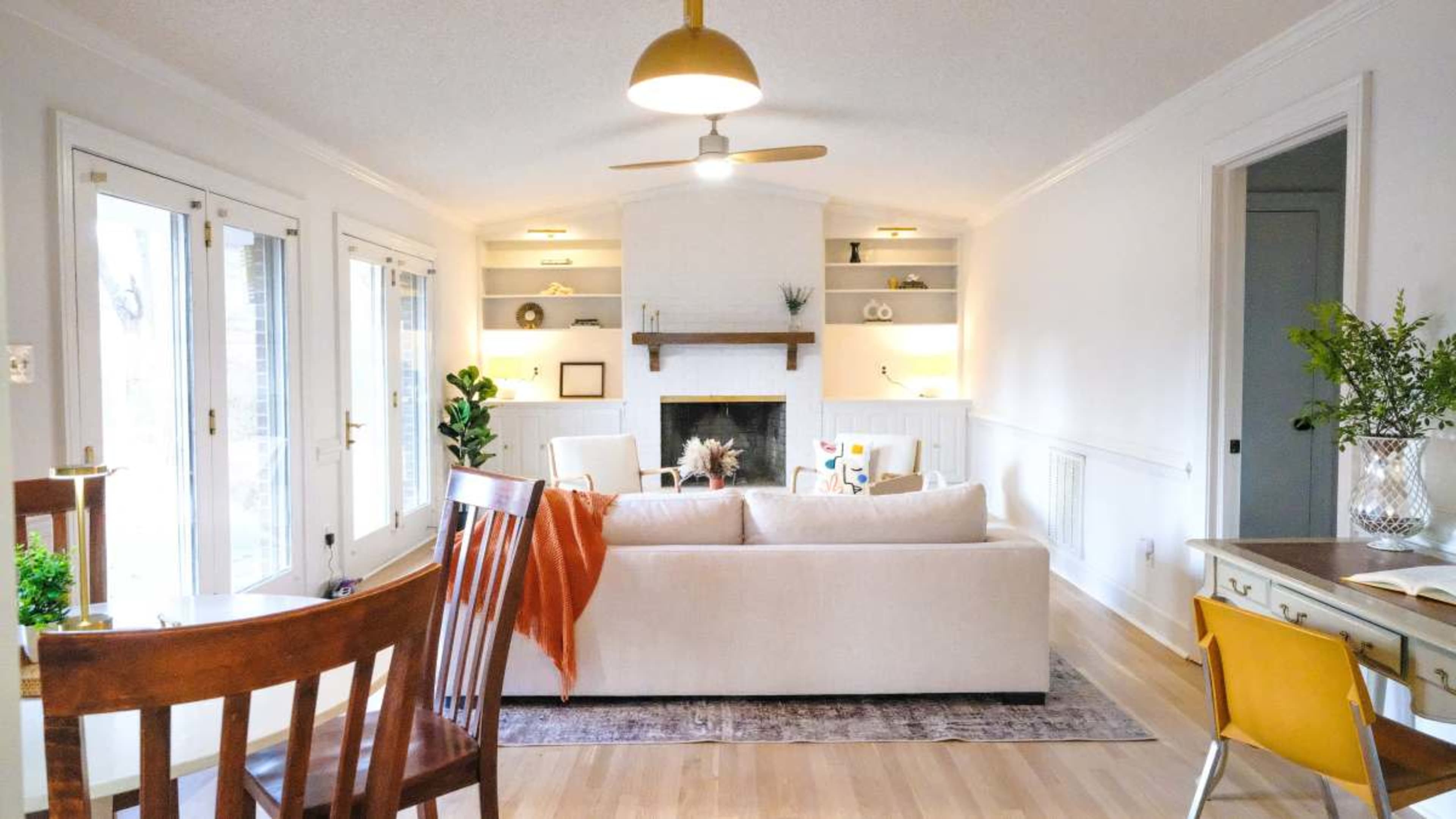 The image shows a light and airy living room with a beige sofa, a fireplace, and built-in shelves, along with a dining area featuring wooden chairs and a small table.