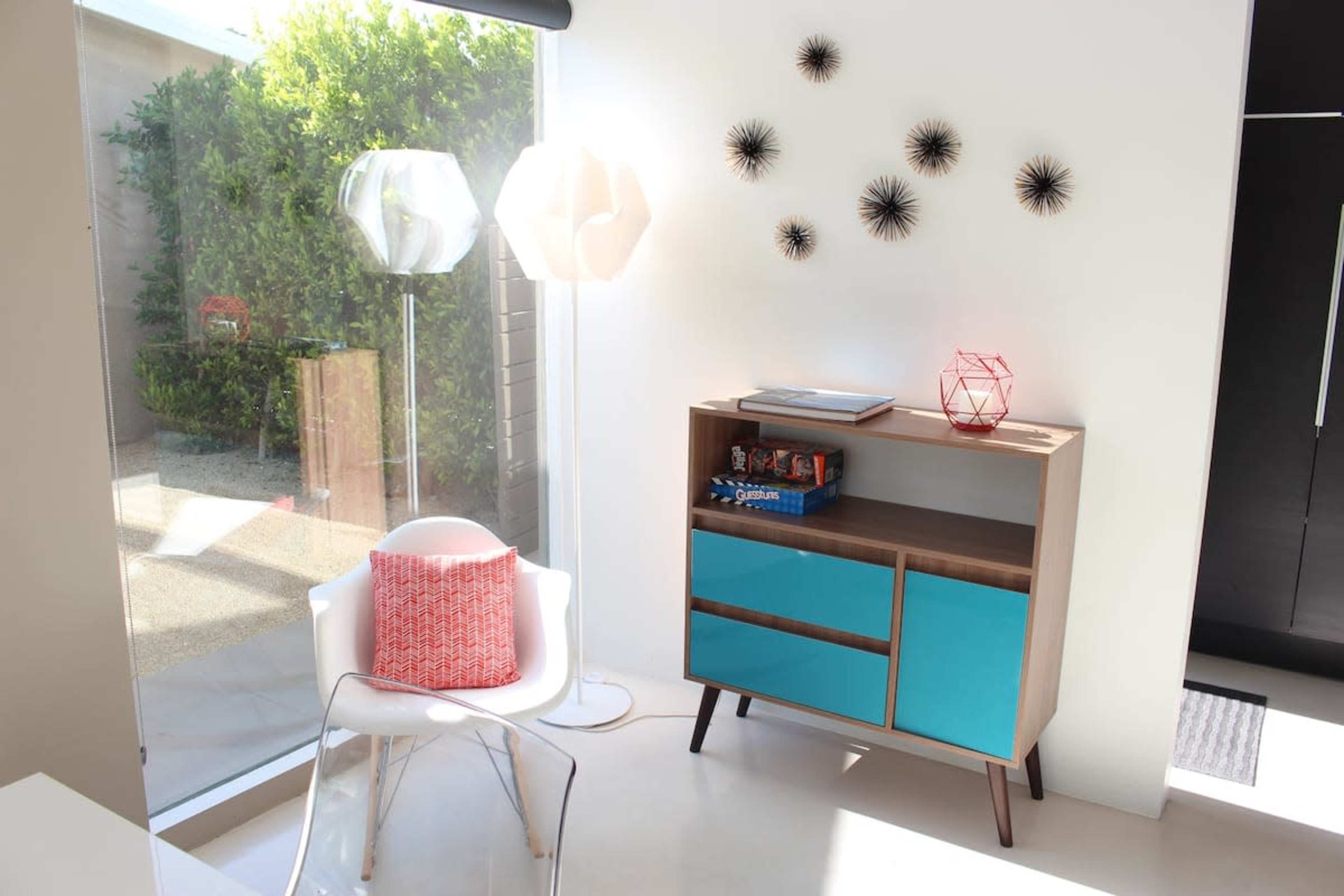 The image shows a bright room featuring a modern turquoise cabinet, a white chair with a red cushion, decorative wall accents, and floor lamps against a large window.