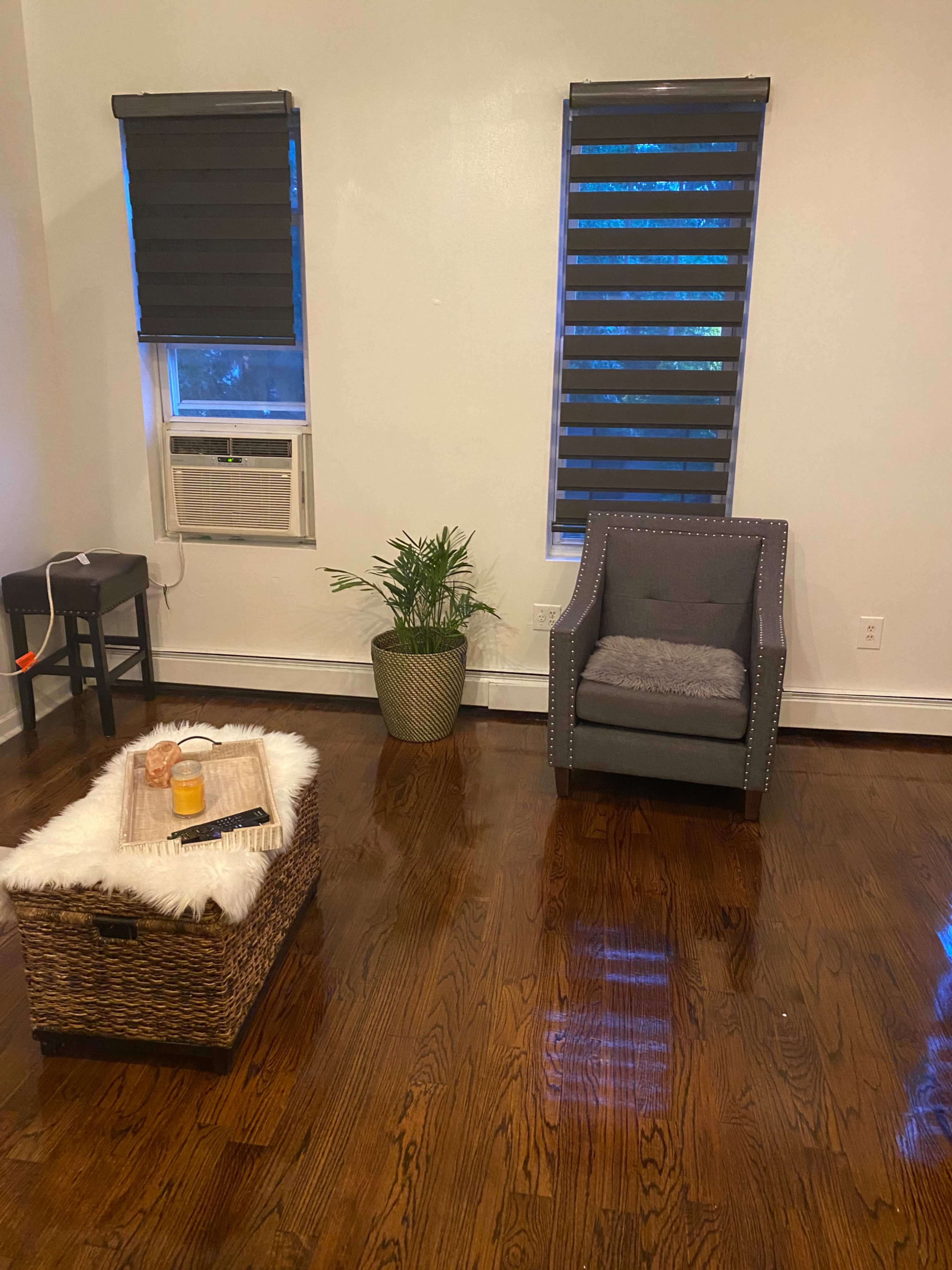 A living room featuring a gray chair, a small table with drinks on it, a woven basket table, and two windows with black blinds.
