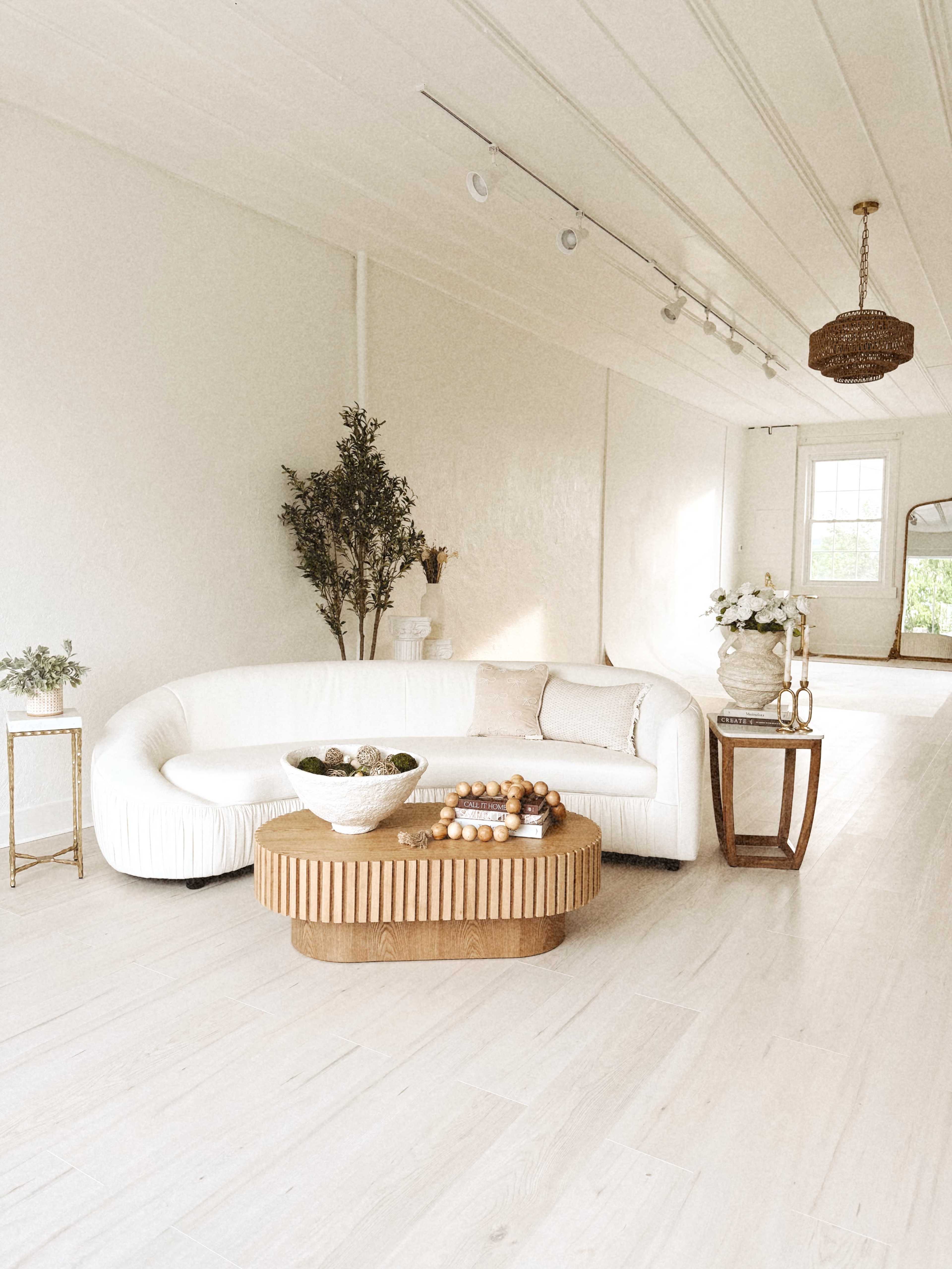 The image shows a bright, minimalist interior featuring a curved white sofa, a wooden coffee table, and decorative elements like plants and vases.