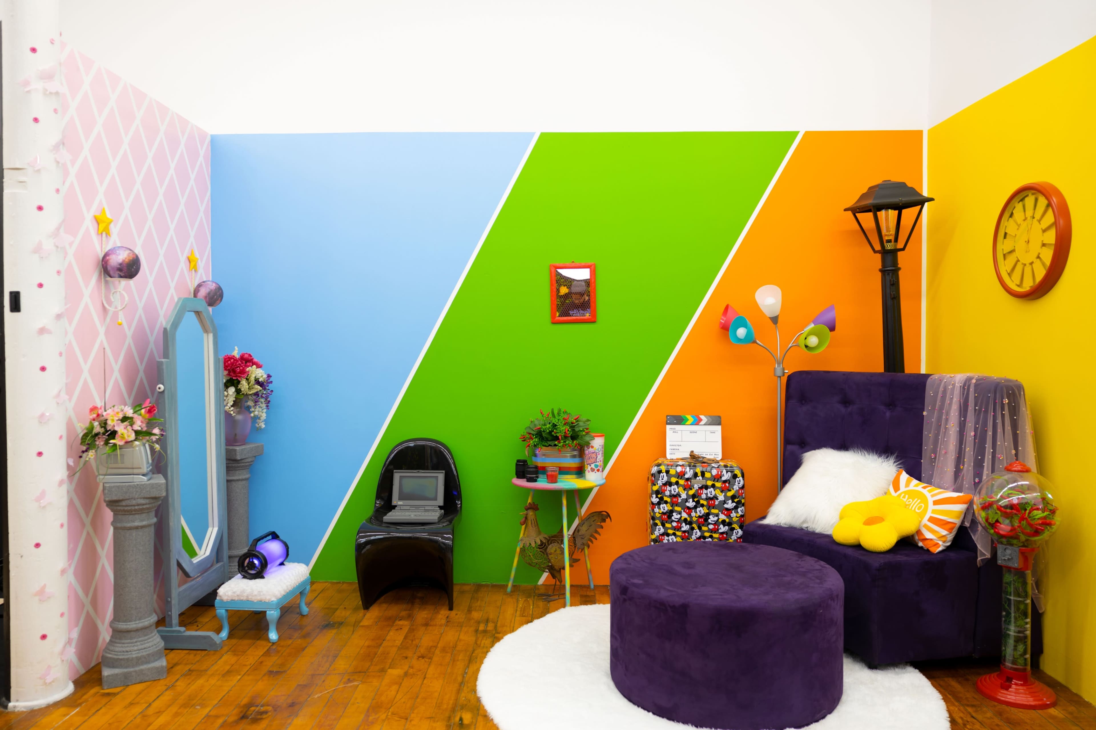 A vibrant room with colorful walls in pink, blue, green, and orange, featuring a purple chair, a round purple ottoman, a large mirror, decorative plants, and a lamp.