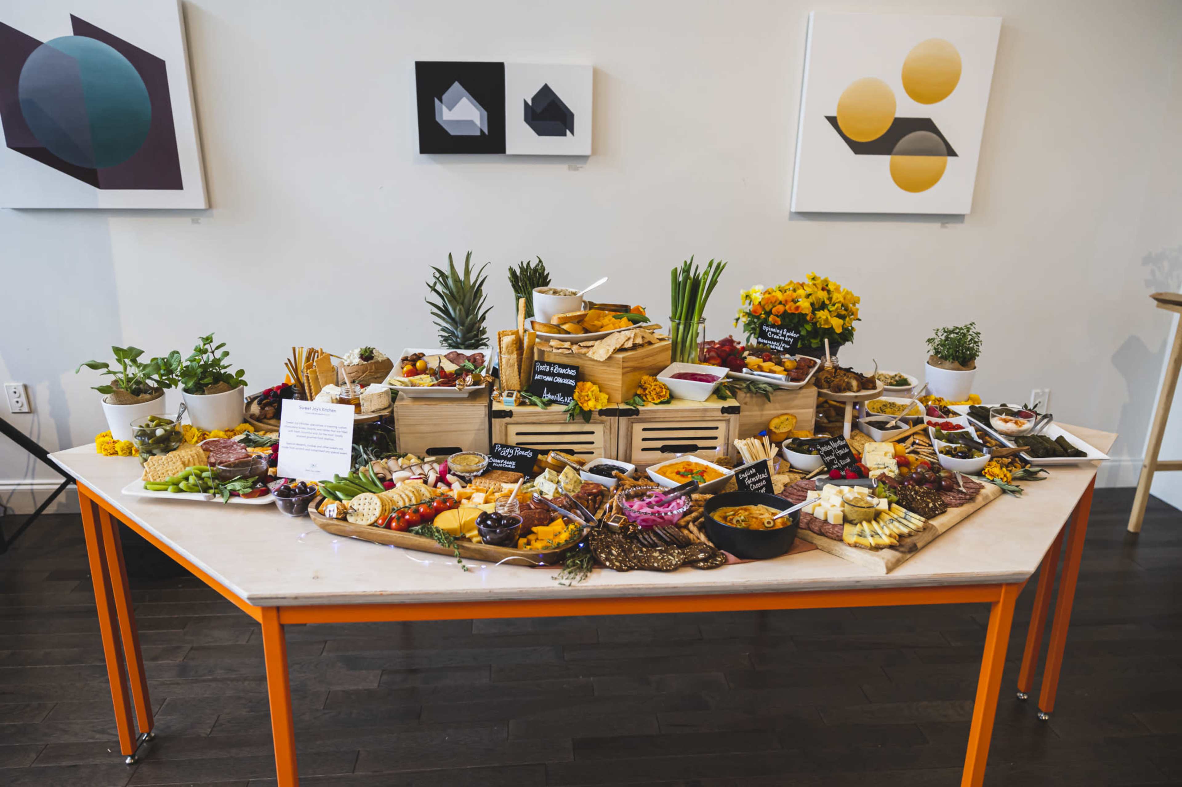 A large, arranged table features a variety of foods, including fruits, vegetables, cheeses, and snacks, along with decorative elements like plants and flowers.