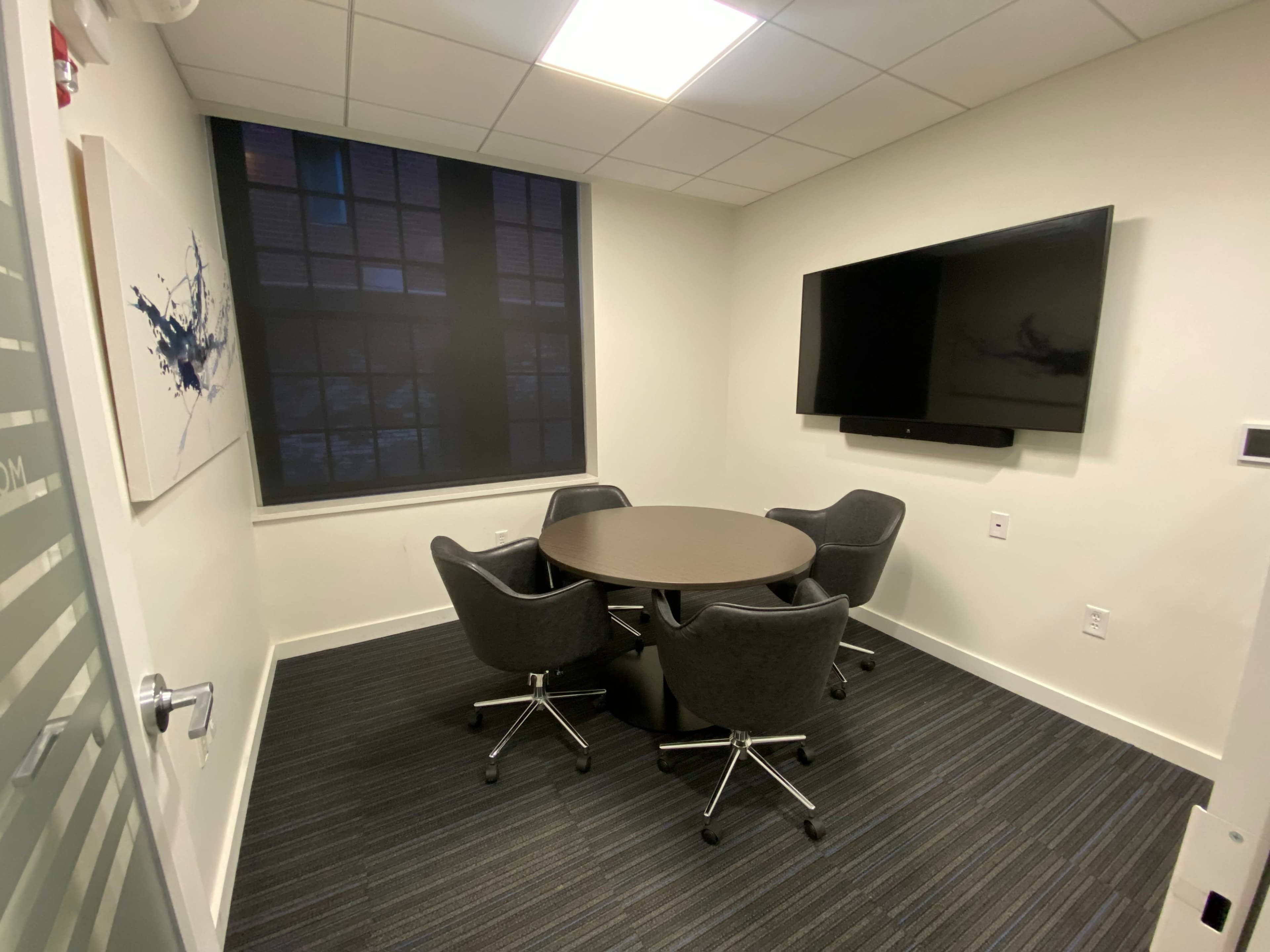 A small meeting room featuring a circular table surrounded by four chairs, with a large wall-mounted television and a window.