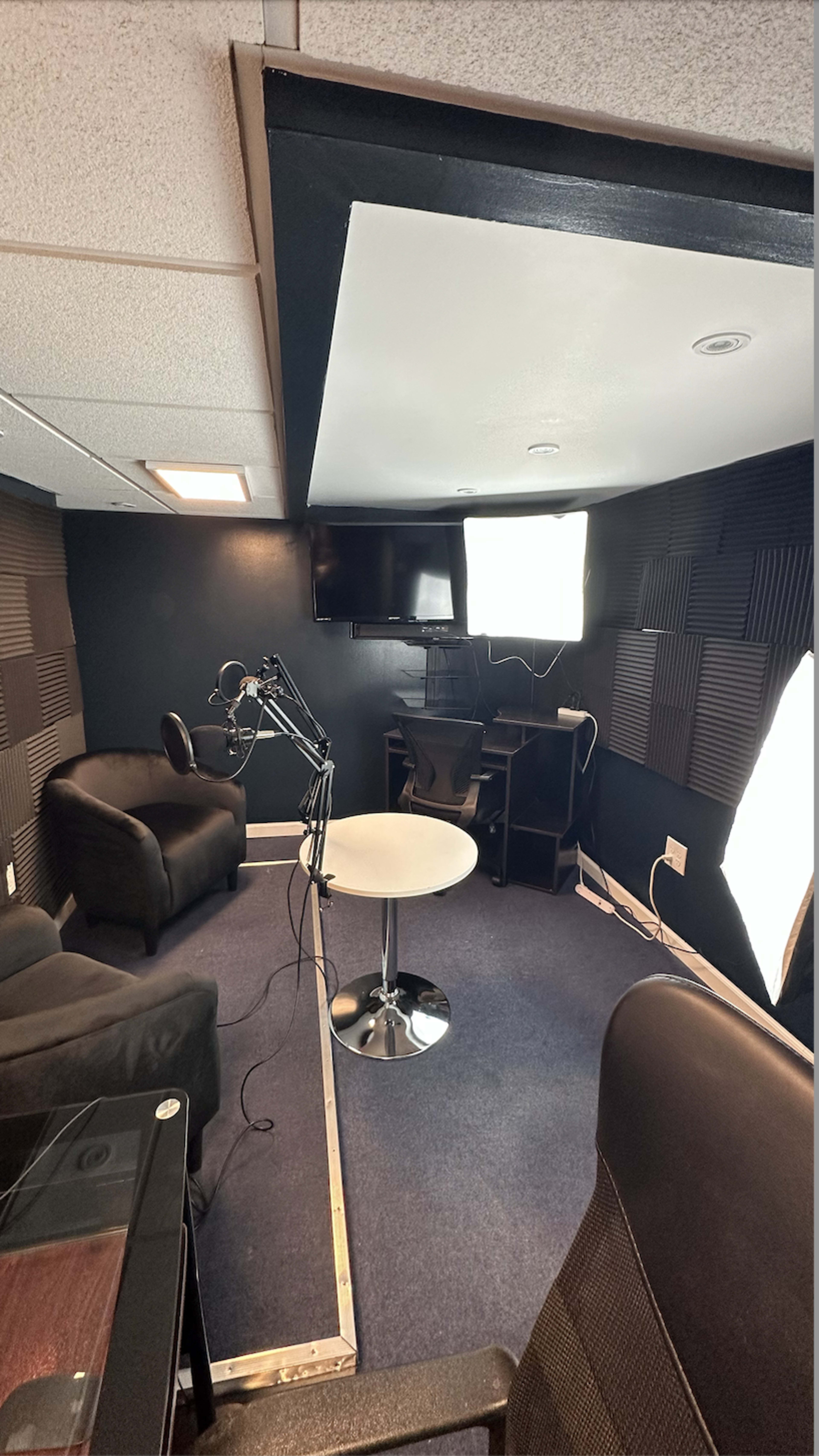 A small recording studio featuring two black chairs, a round table, a microphone on a stand, and softbox lights in front of a wall-mounted television.
