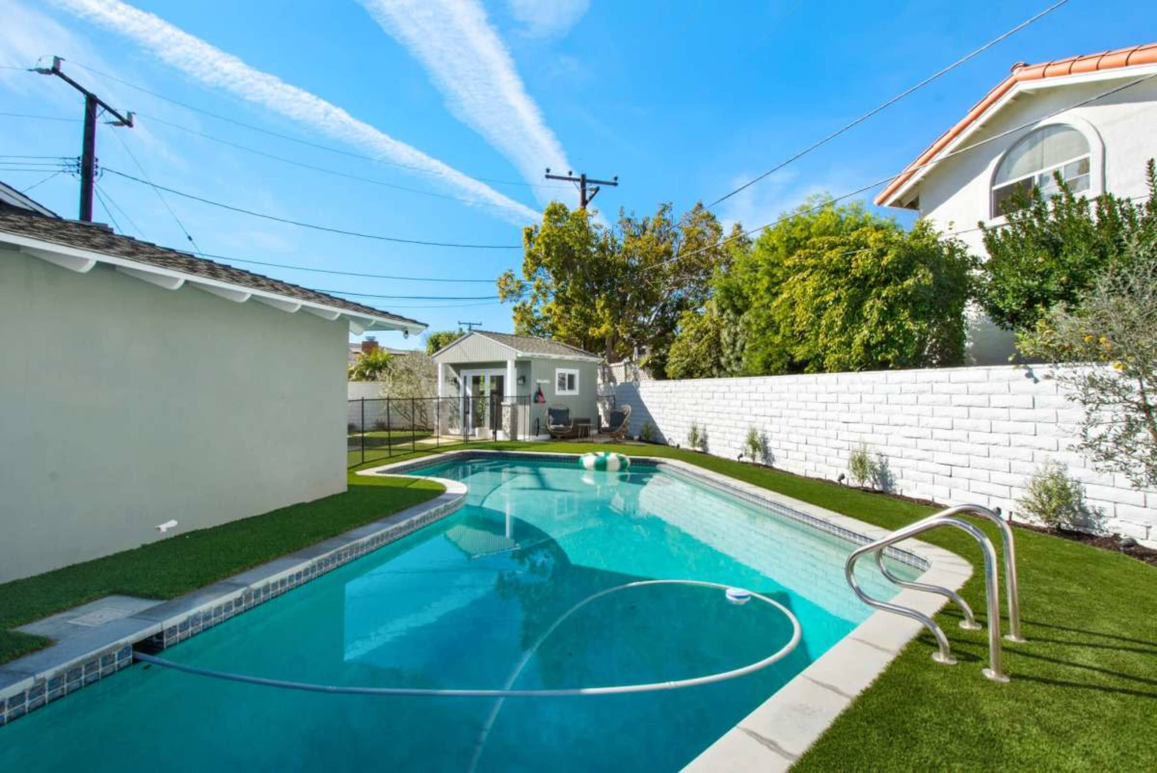 A rectangular swimming pool is surrounded by green grass and bordered by a white wall, with a small building visible in the background under a clear blue sky.