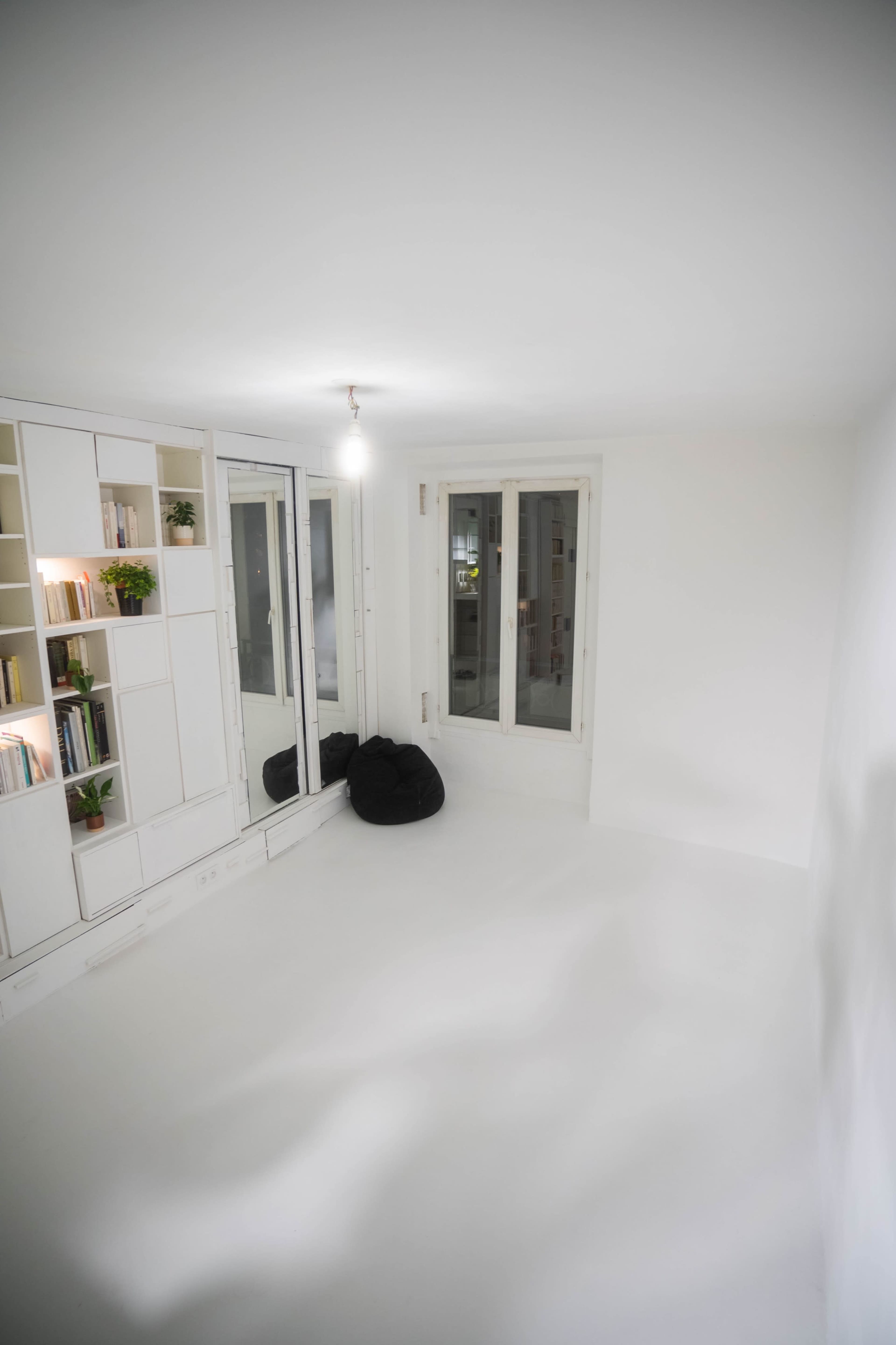 The image shows a minimalist room with white walls, a mirrored closet, shelving filled with books, and two black bean bags on a white floor.