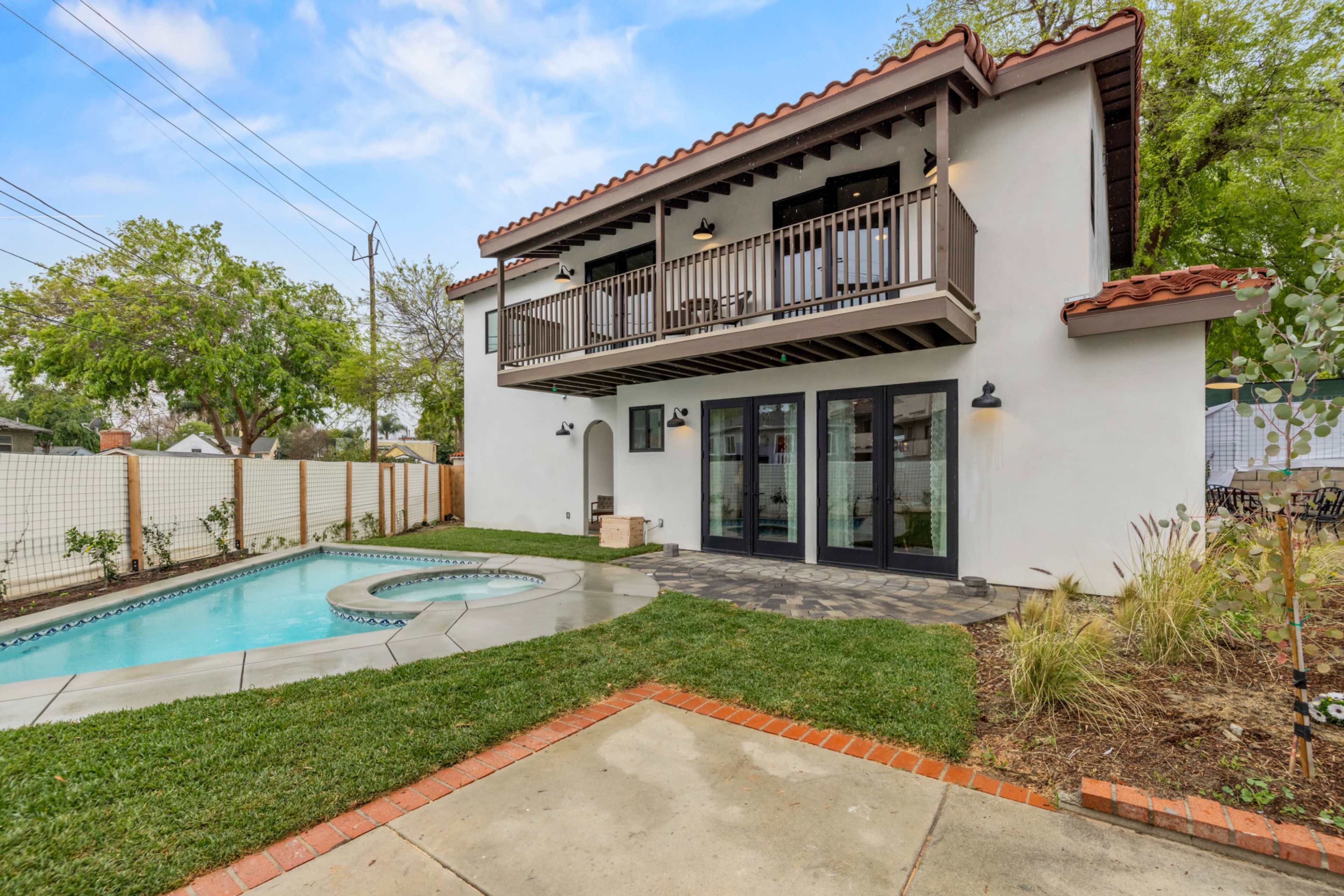 A two-story house with a tiled roof features a balcony, large windows, and a swimming pool in the backyard surrounded by grass and landscaping.