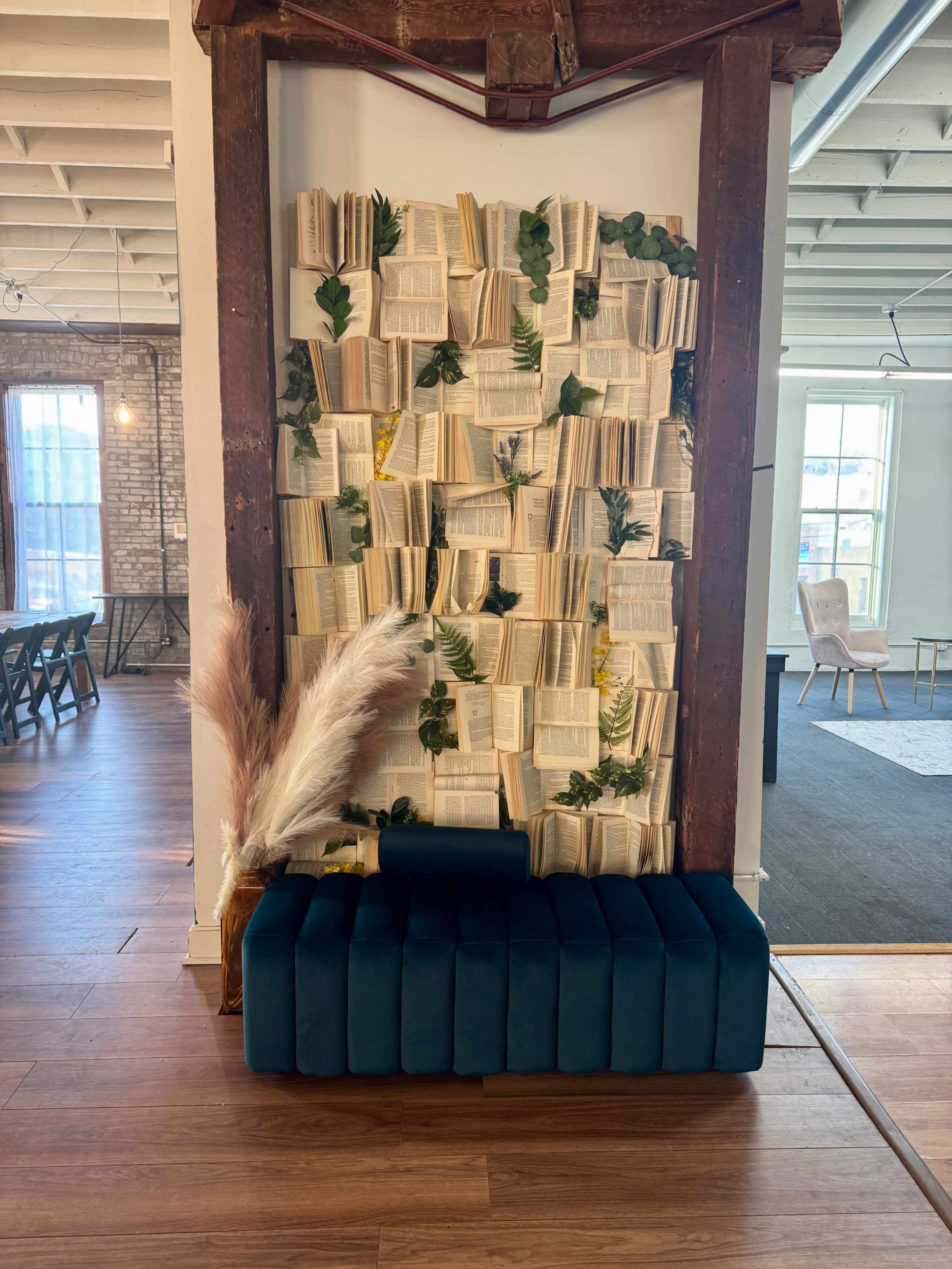 The image shows a decorative wall made of arranged book pages adorned with greenery, beside a modern teal bench in an open space with wooden flooring.