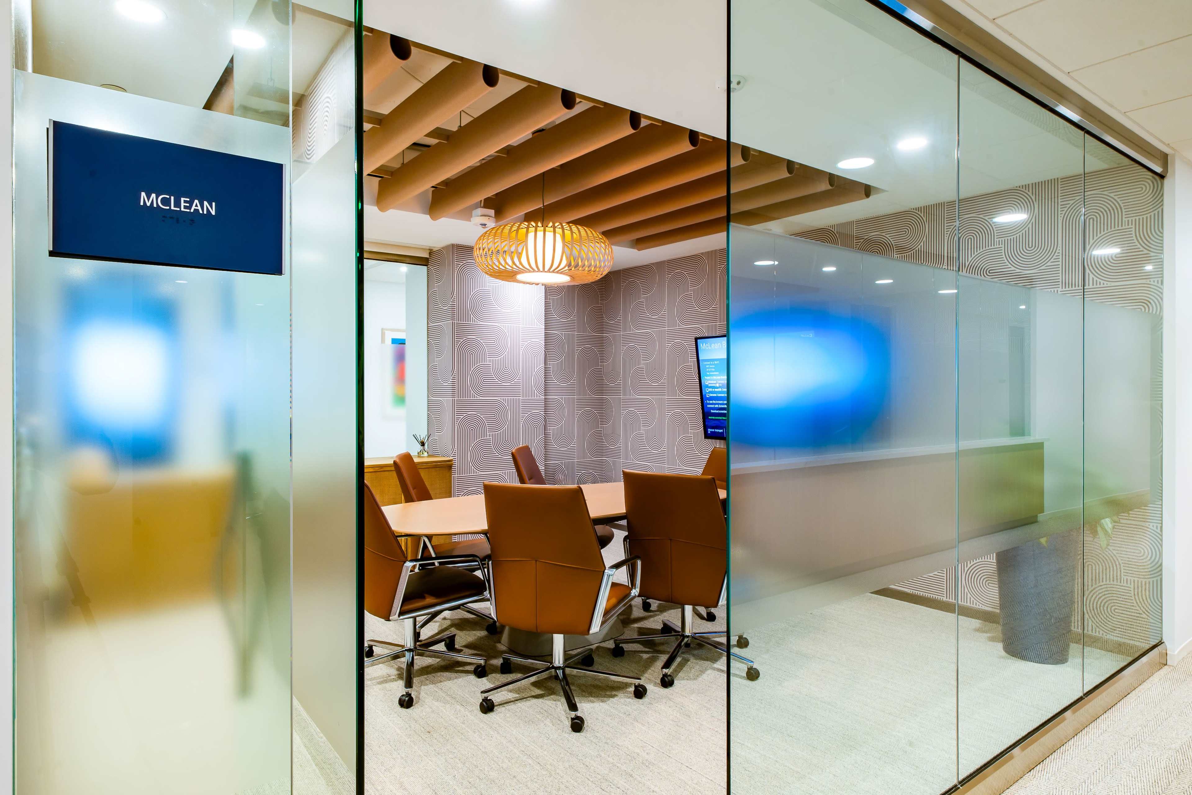 The image shows a modern conference room with a glass wall, featuring a wooden table surrounded by brown office chairs and decorative lighting.