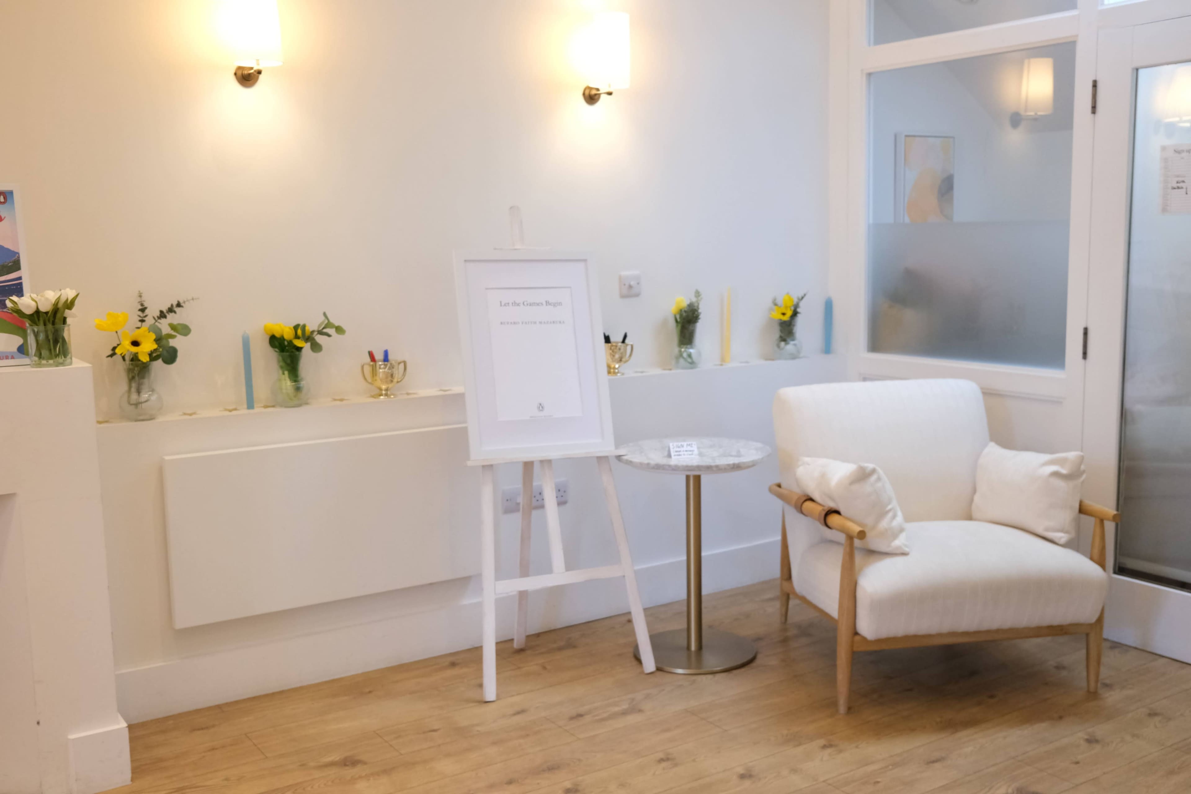 The image shows a tidy waiting area with a white chair beside a small table, decorated with flowers and a sign.