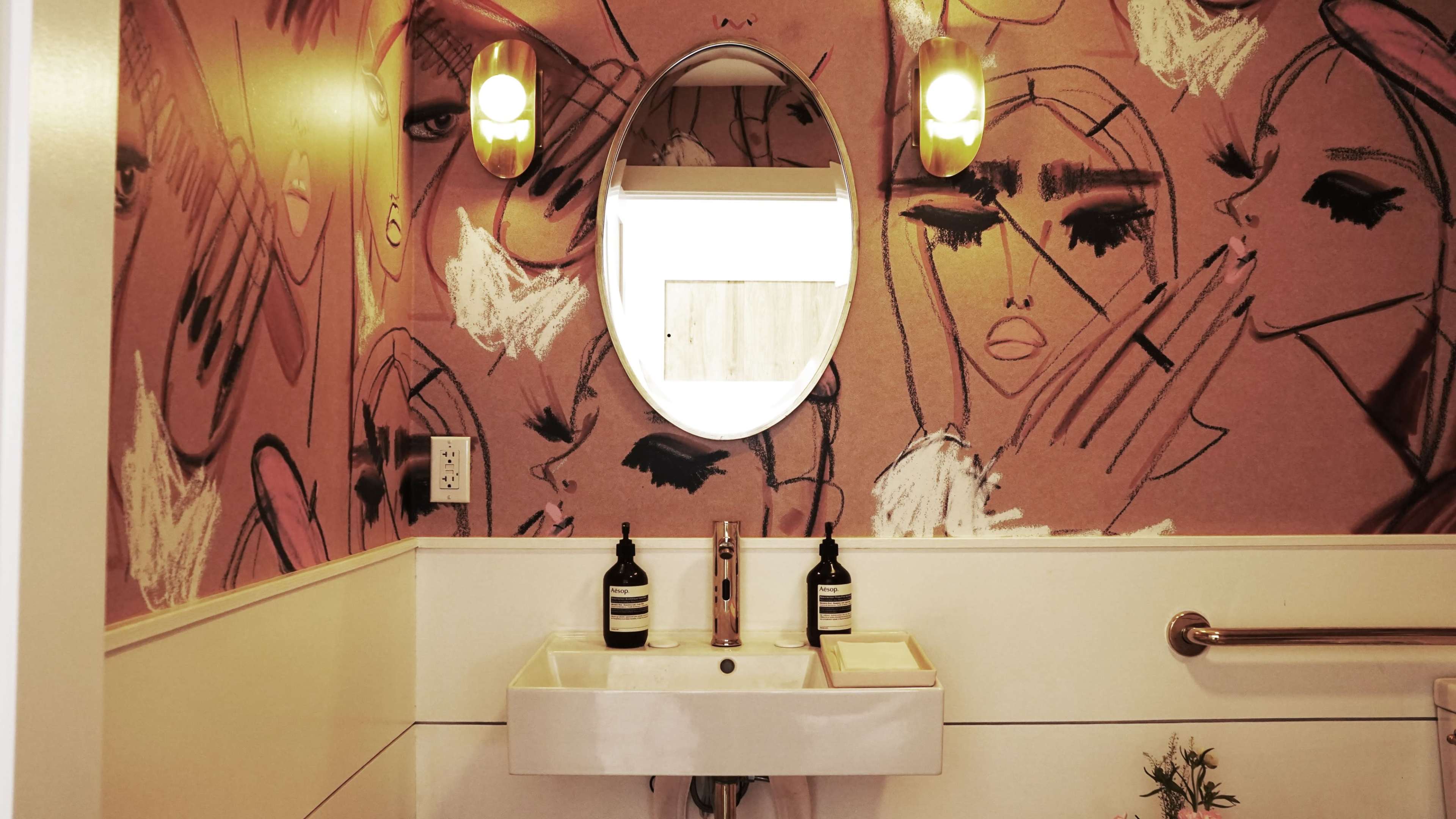 The image shows a bathroom featuring a modern sink, oval mirror, and walls adorned with artistic, graphic patterns of faces.