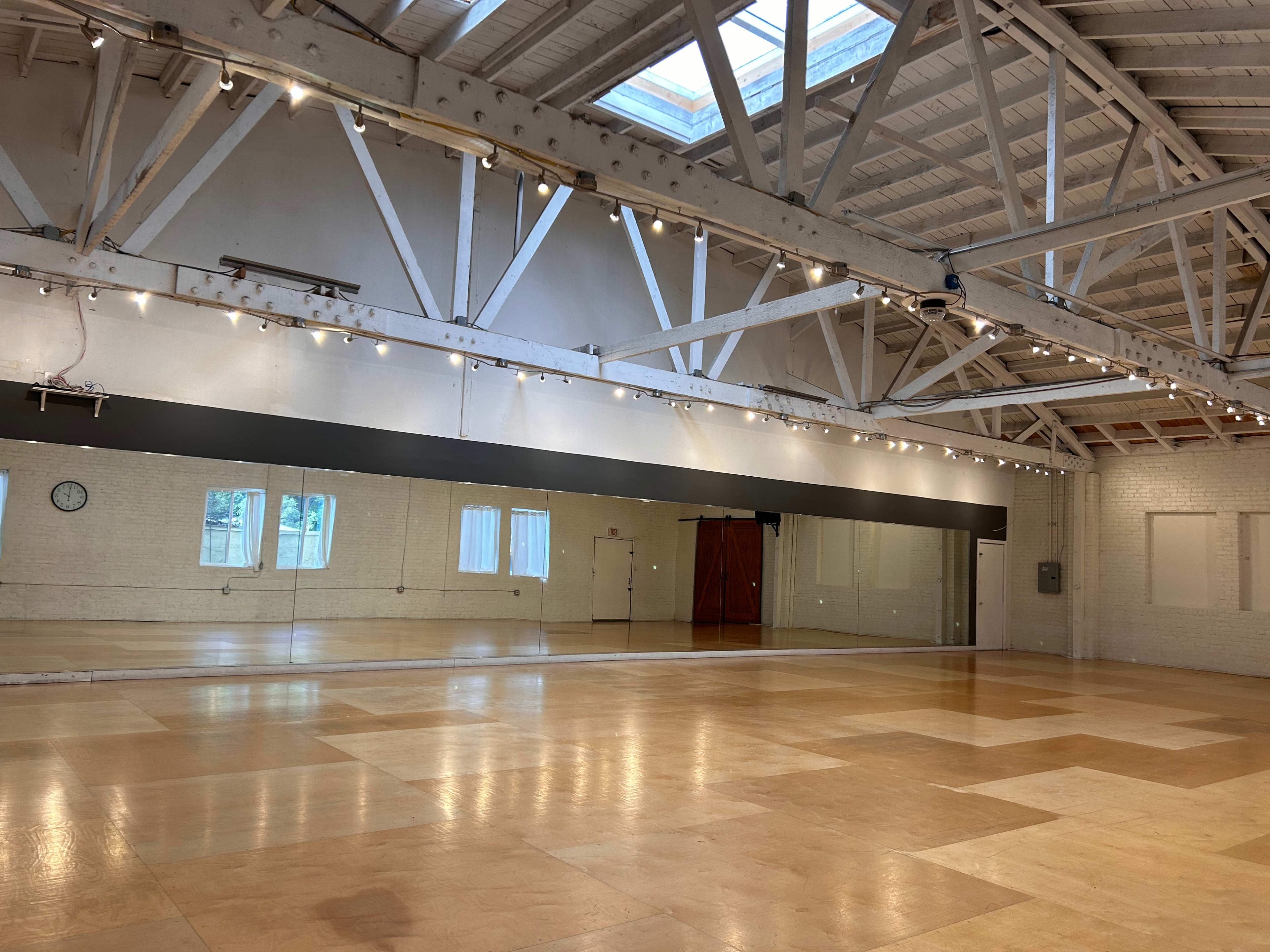 An empty dance studio features polished wooden floors, large mirrors on one wall, and a skylight above.