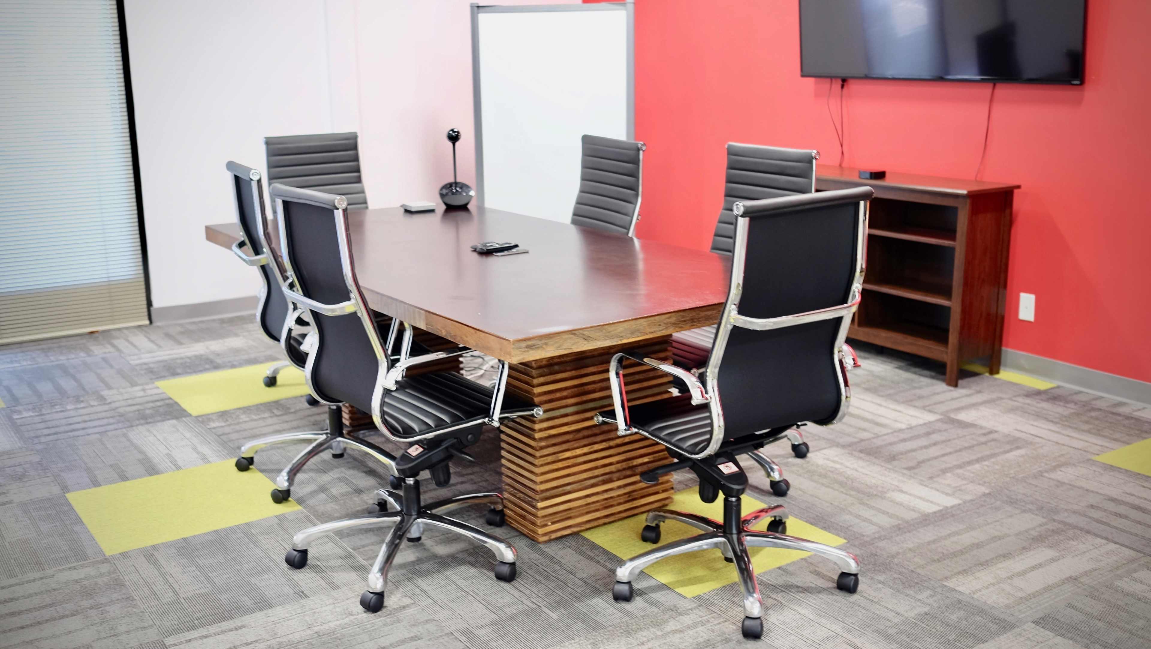 A modern conference room features a rectangular wooden table surrounded by six black office chairs, with a television mounted on the wall and a whiteboard in the corner.