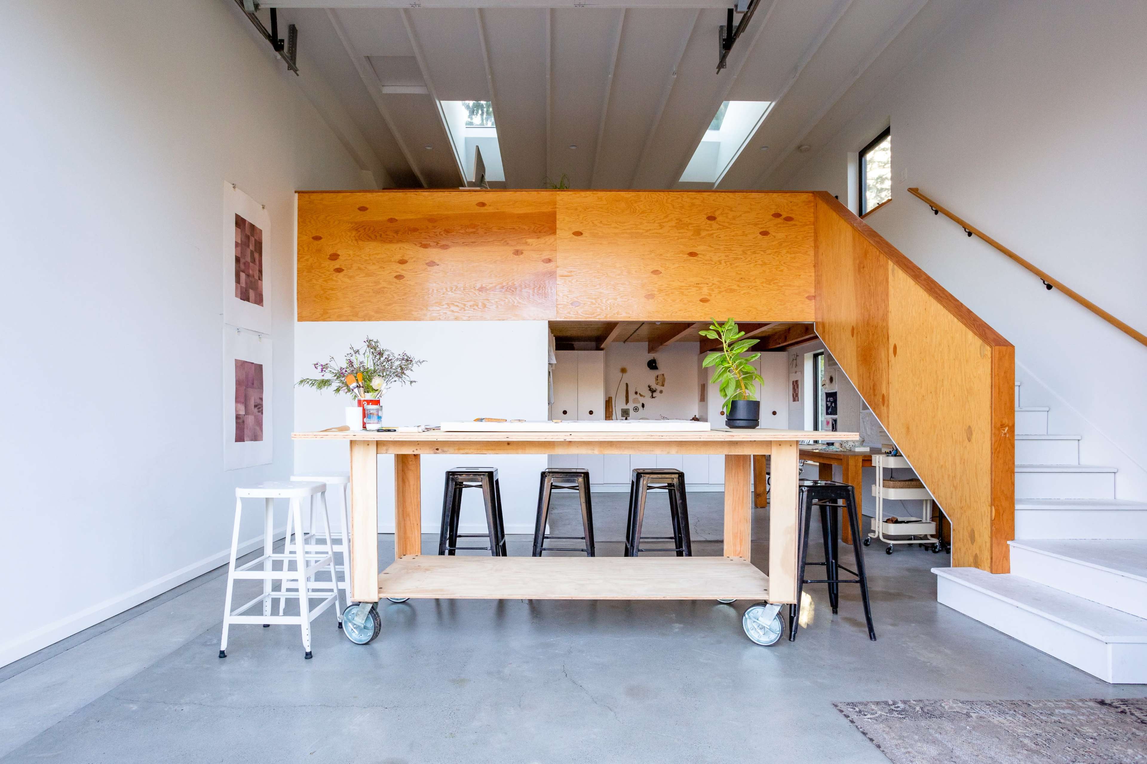 A modern kitchen area with a wooden island on wheels, black metal stools, and a staircase in the background.