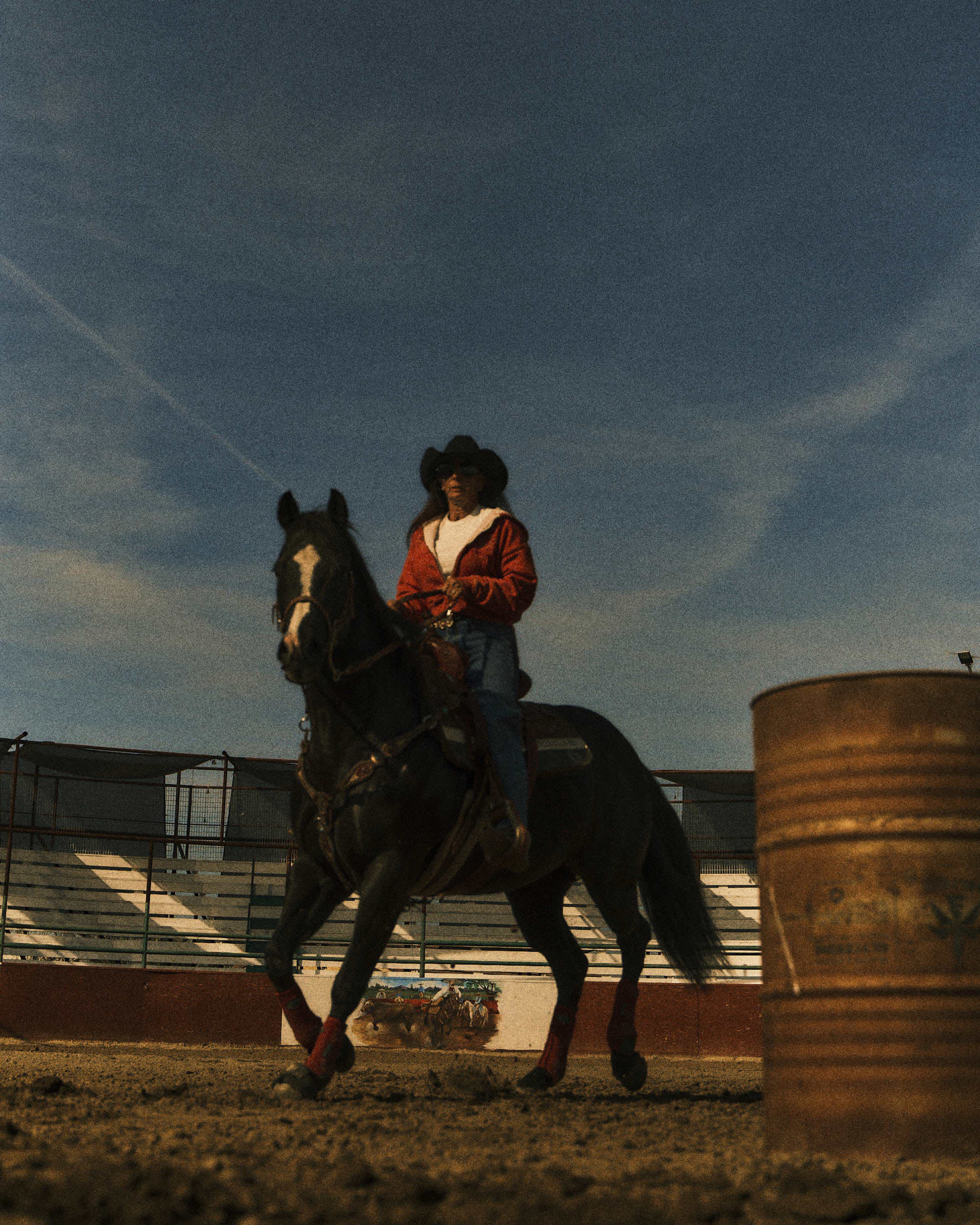 A person wearing a cowboy hat and a red jacket rides a black horse around a rodeo arena, with a metal barrel in the foreground.