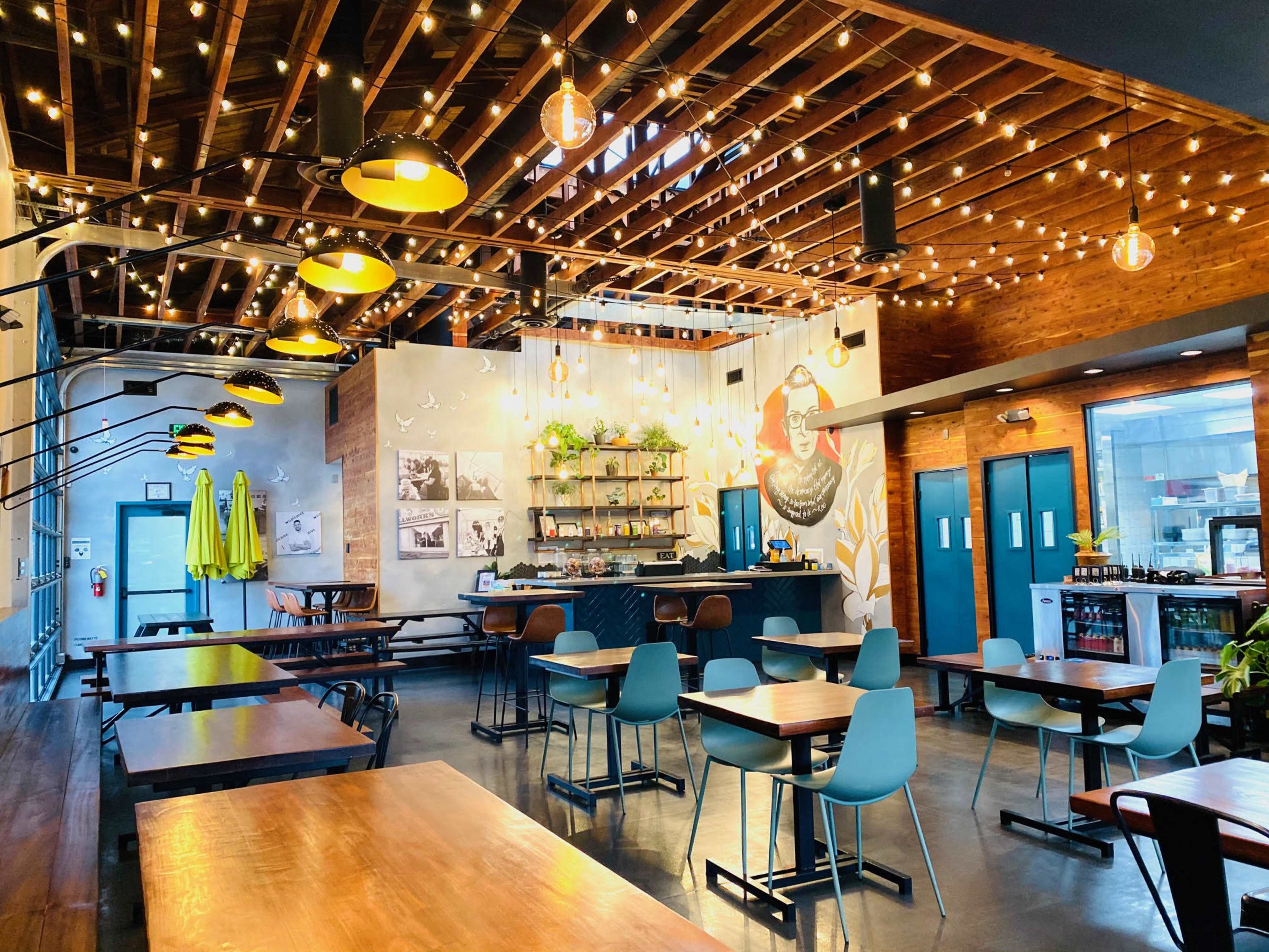 A modern restaurant interior with wooden tables, colorful chairs, string lights overhead, and a bar area adorned with plants and artwork.