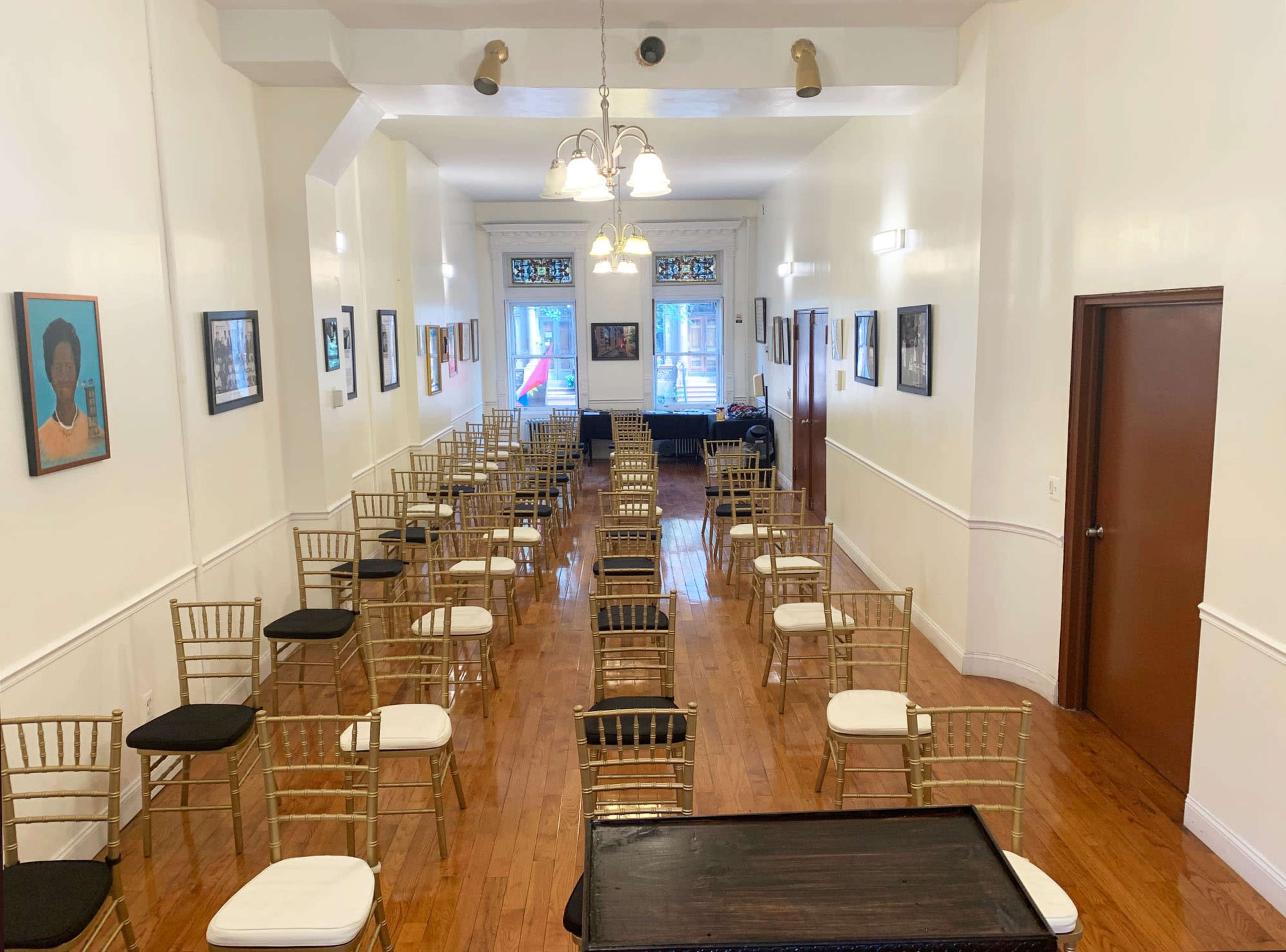A long, narrow room with rows of empty gold chiavari chairs and framed artwork on the walls.