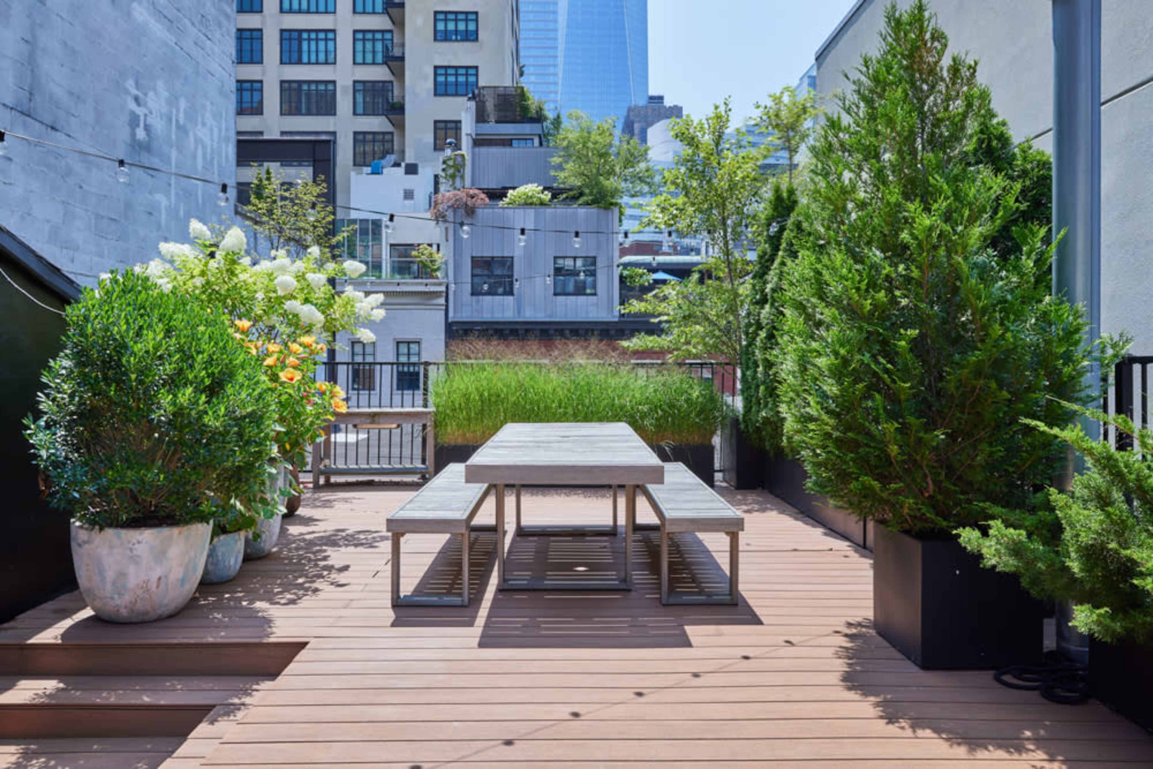 A rooftop terrace featuring wooden decking, a table with benches, and large planters filled with greenery.