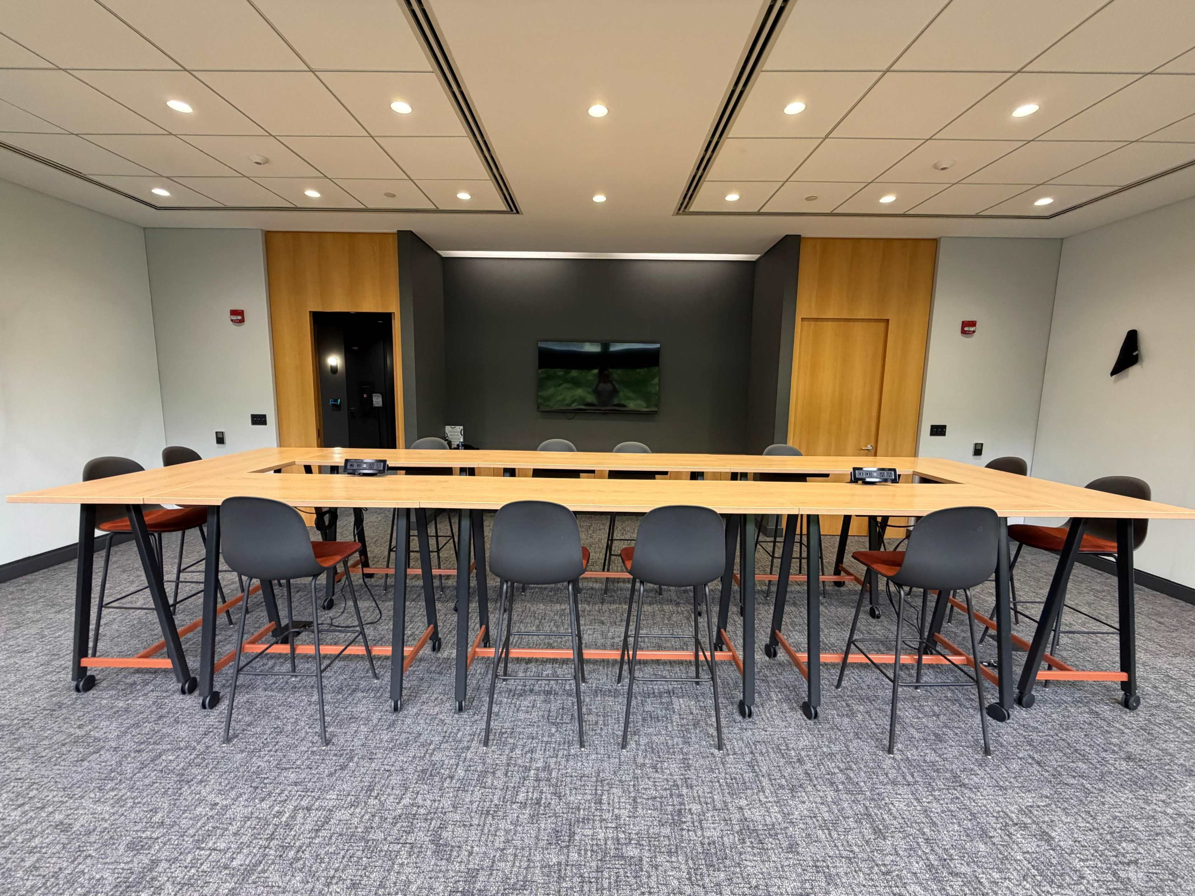 A long, rectangular conference table is surrounded by black and orange chairs in a modern meeting room with a large screen on the wall.