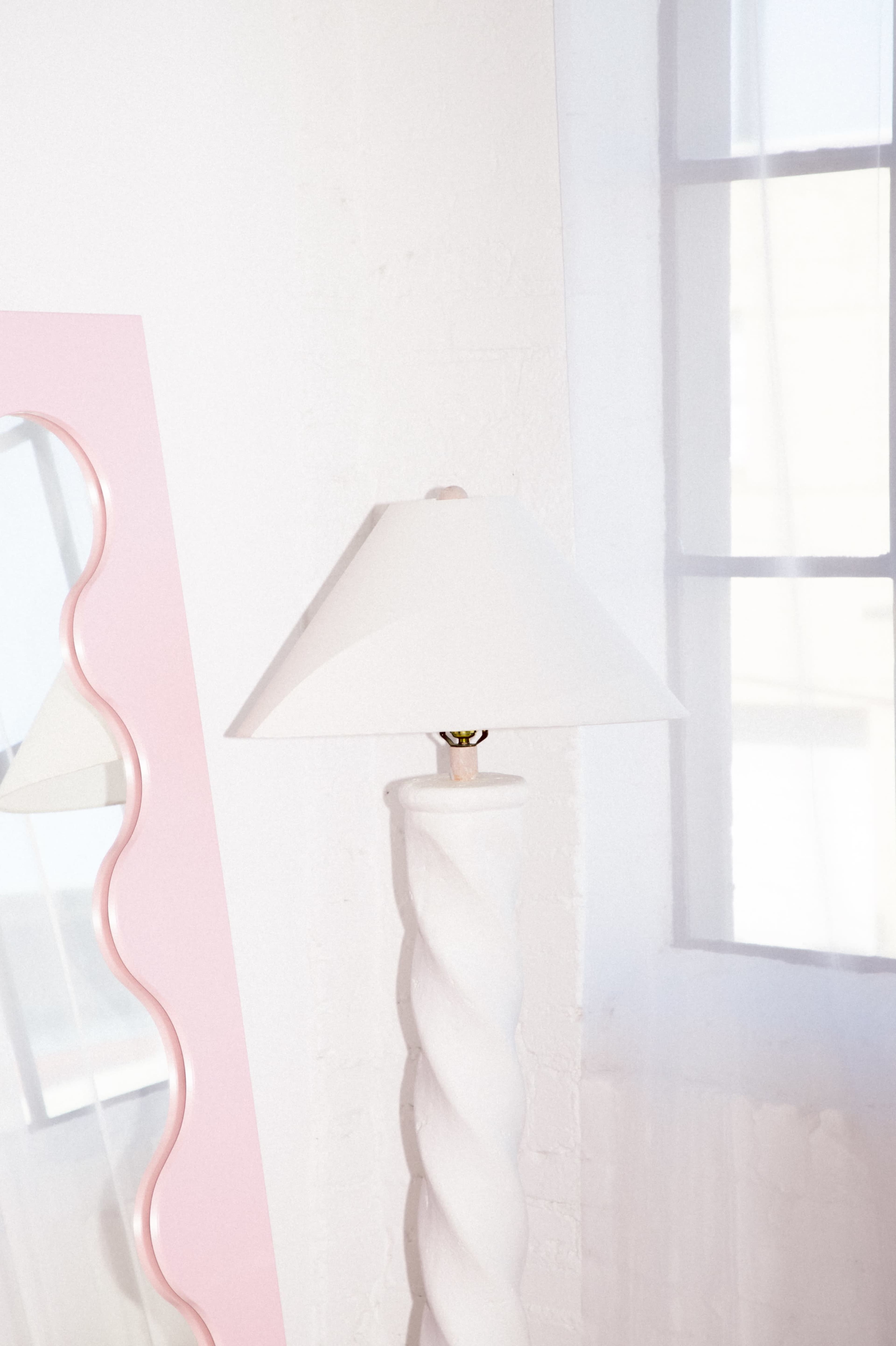 A white lamp with a spiral base stands next to a pink-framed mirror near a window.