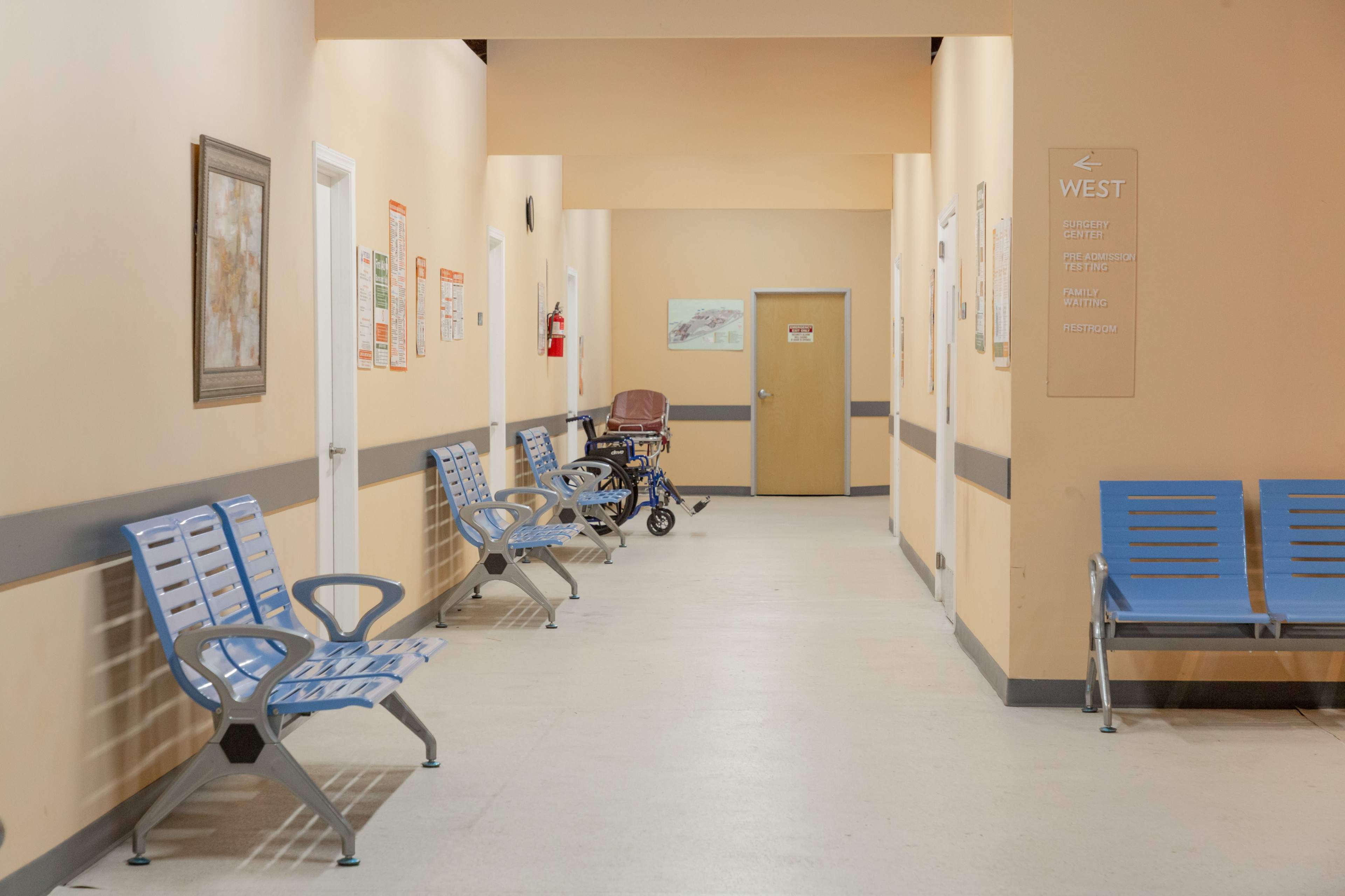 A hospital hallway lined with blue benches and a baby stroller, with doors and signs along the walls.