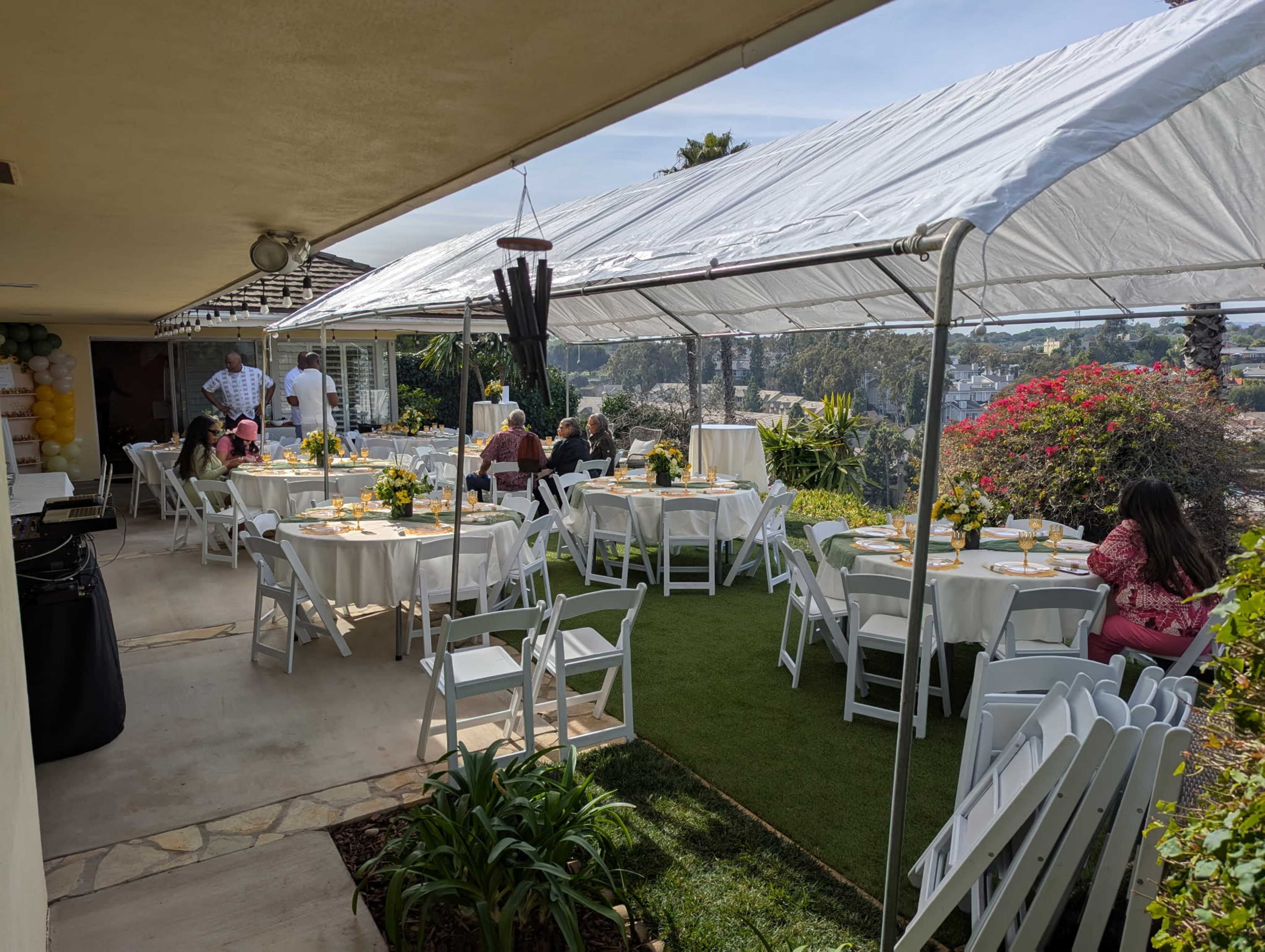 A spacious outdoor setting features several tables dressed in white tablecloths, surrounded by chairs, with a tent overhead and guests enjoying a gathering with a scenic view.