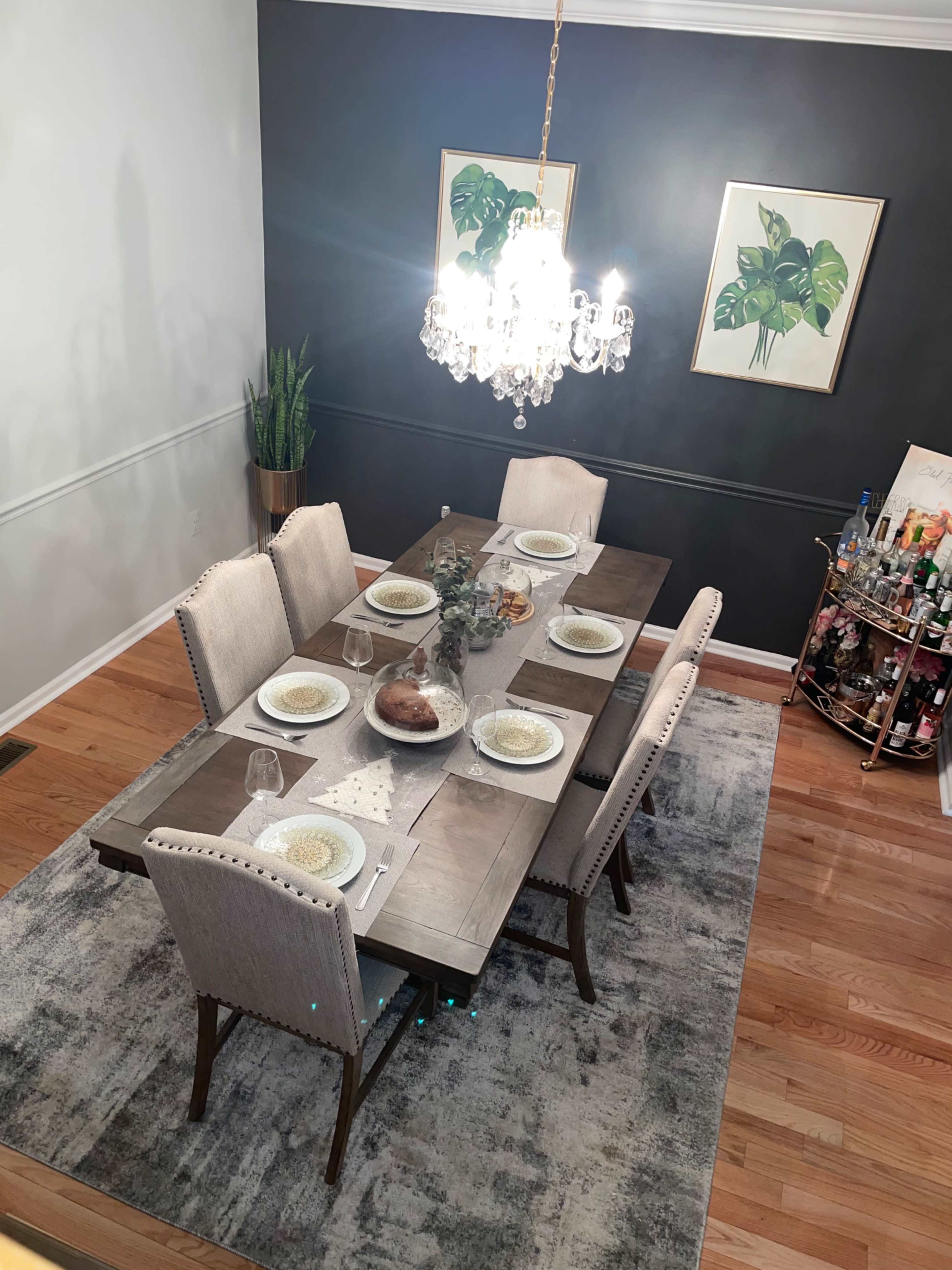 A dining room table is set for a meal, surrounded by upholstered chairs, under a chandelier, with decorative plants and a bar cart in the background.