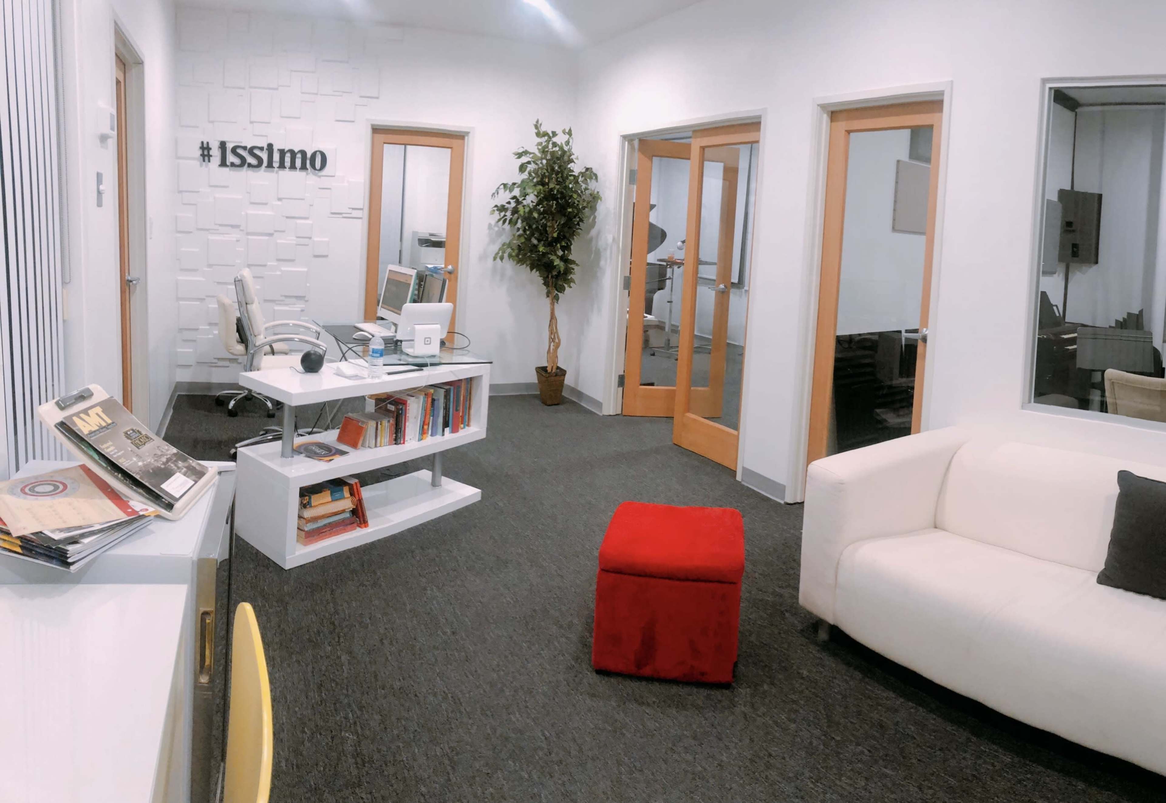 A modern office space featuring a white sofa, a small red ottoman, a desk with books, and a potted plant against a wall with the hashtag "#Isslimo."