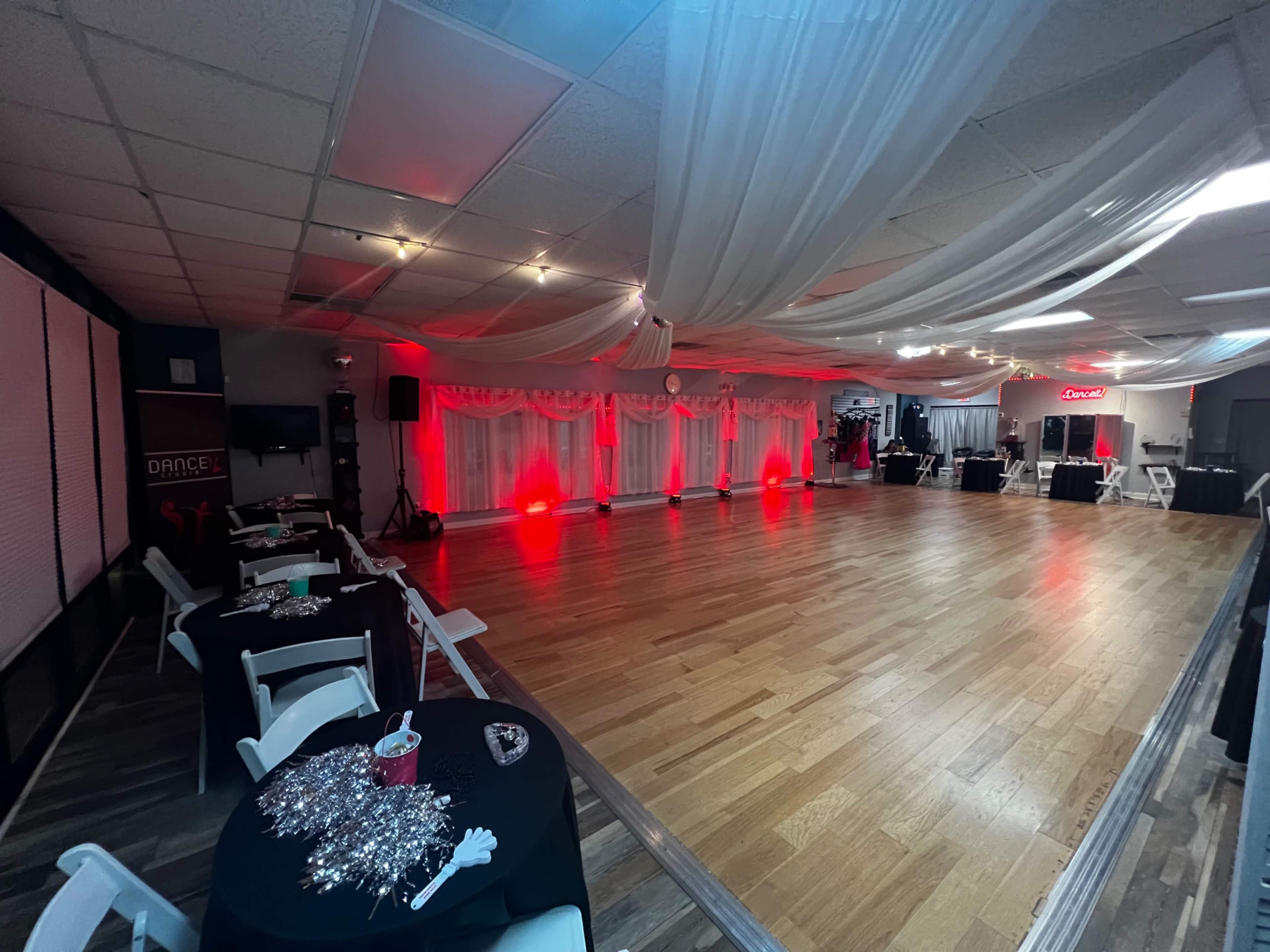 The interior of a dance studio features a spacious wooden floor, decorated with white drapes and red lights, with tables set up along the sides.