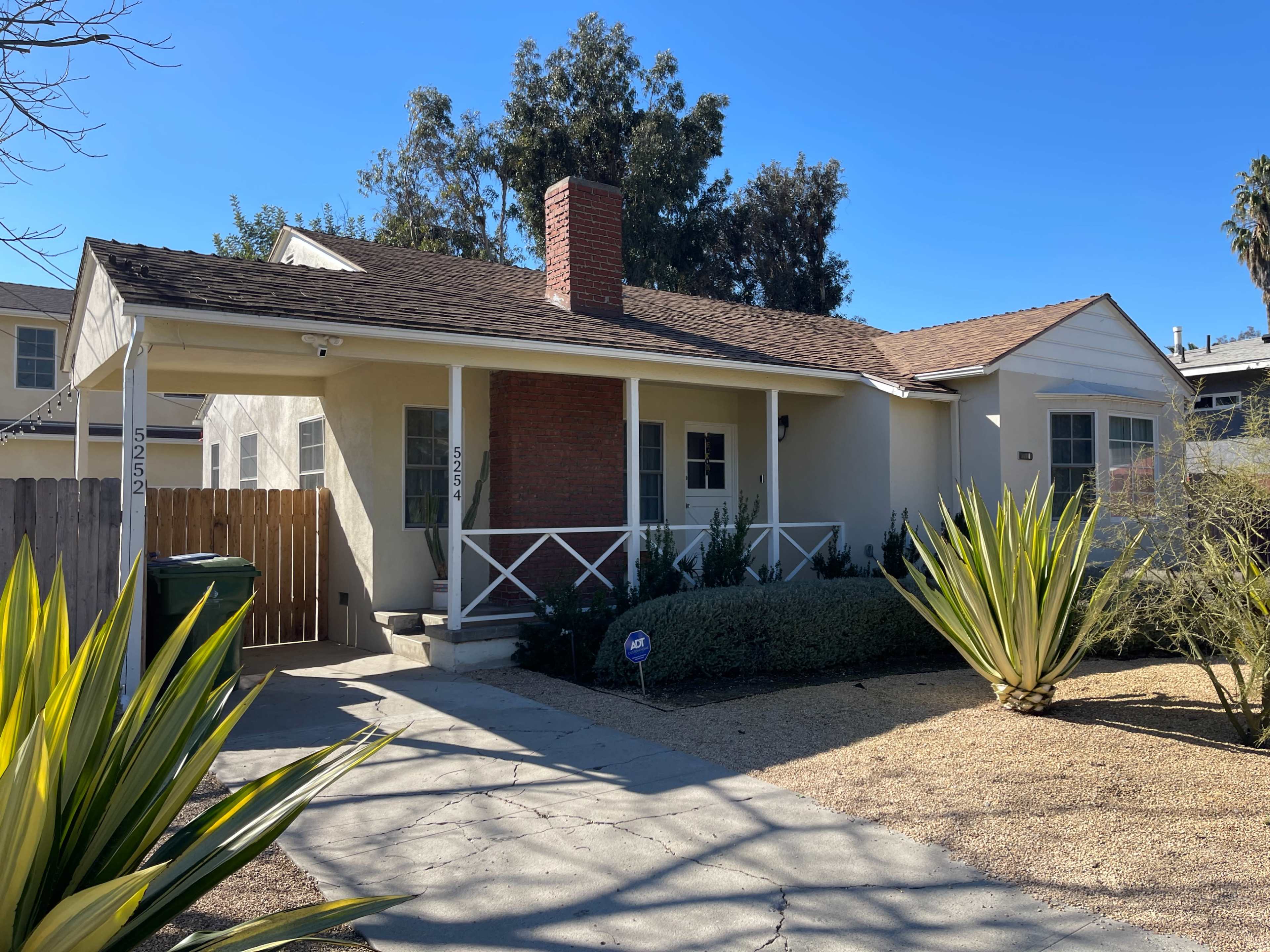 The image shows a single-story house with a front porch, a chimney, and a gravel yard surrounded by low-growing plants.