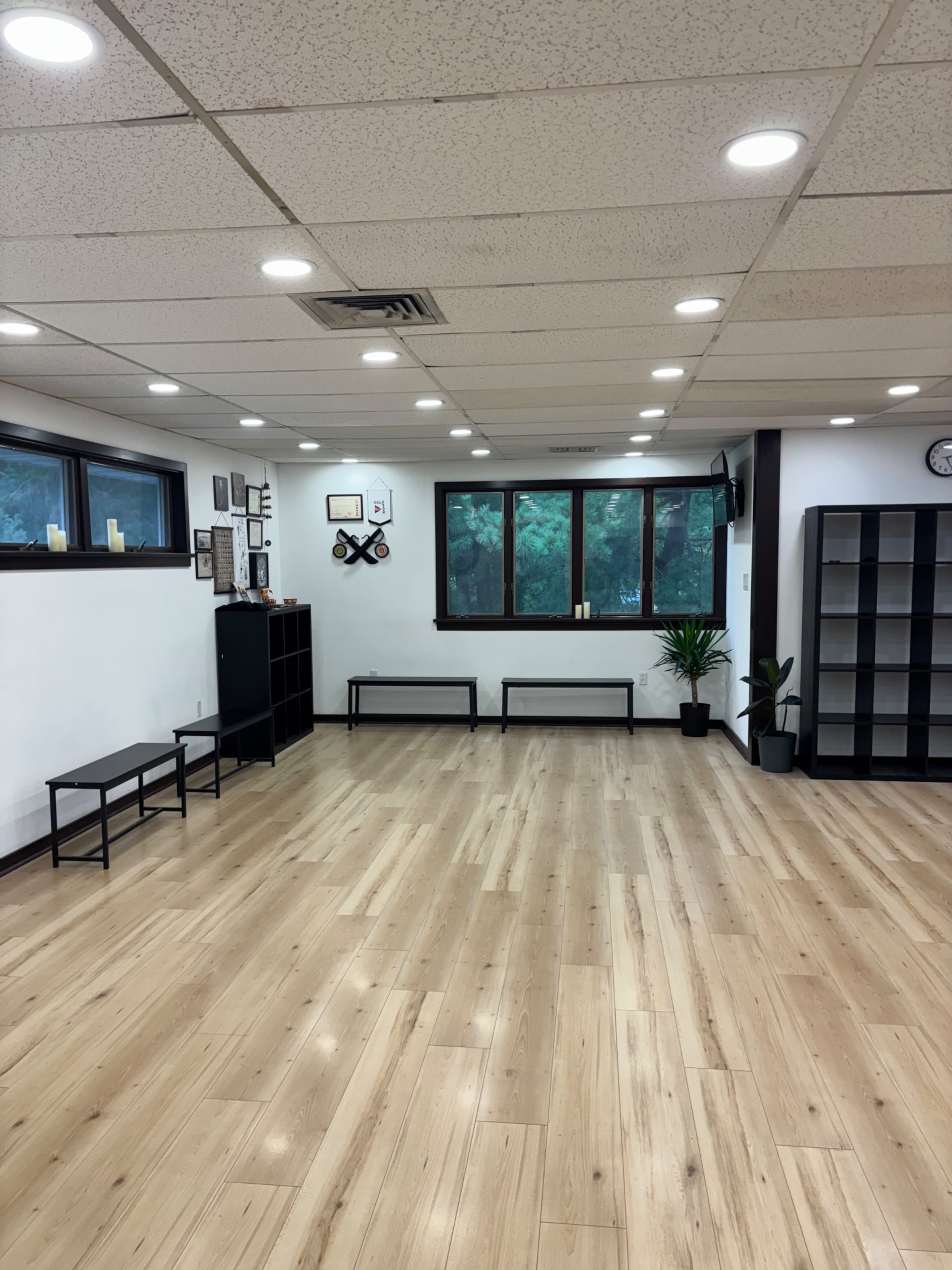 The image shows an empty, well-lit room with wooden flooring and large windows overlooking greenery, furnished with black shelves and benches against the walls.