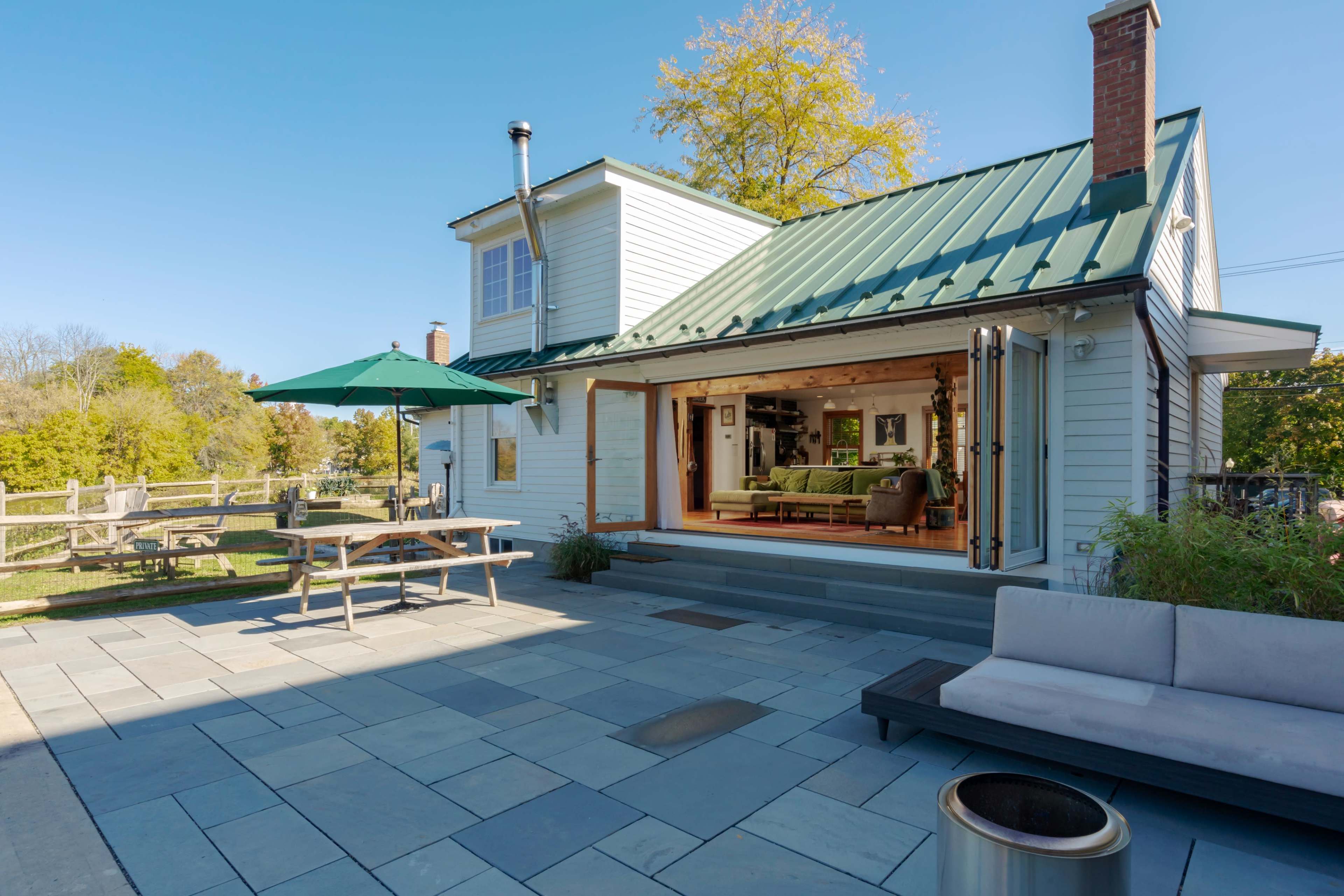 A house with a green metal roof has an open patio area featuring a wooden table, chairs, and an umbrella, leading to a spacious living room with large glass doors.