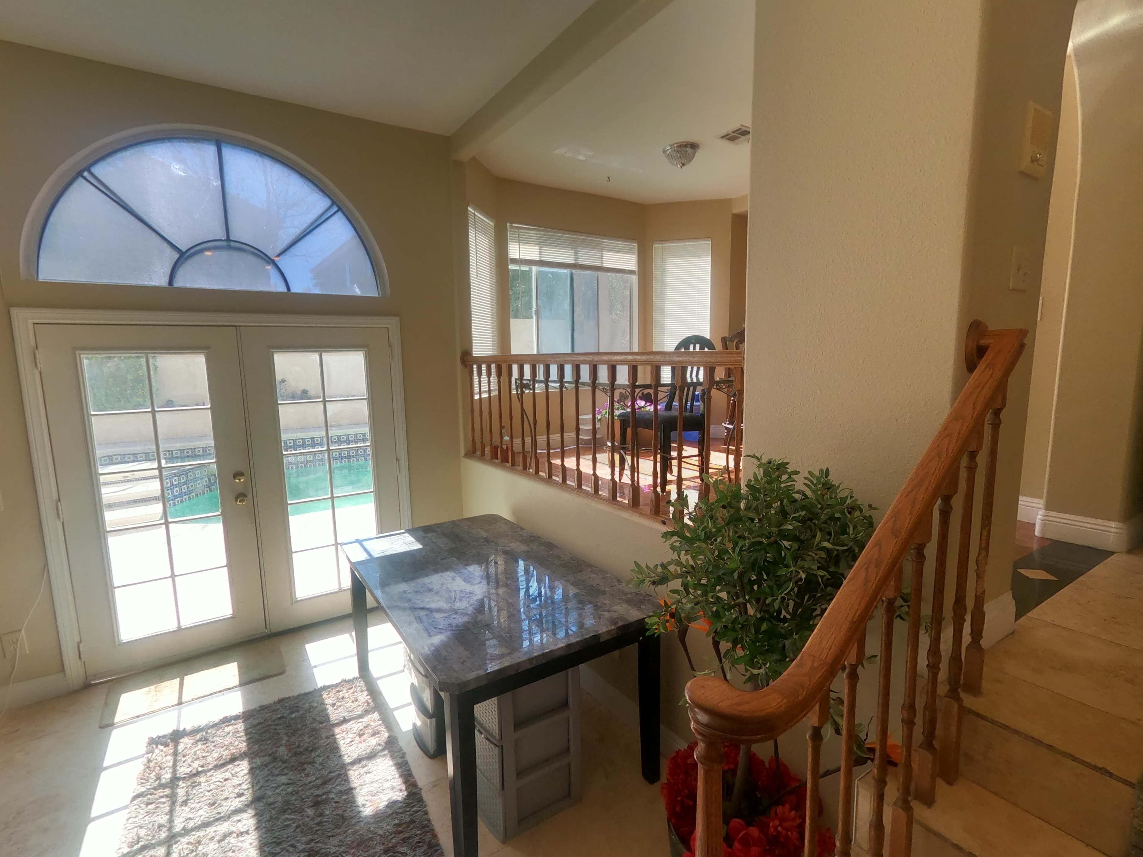 A spacious foyer features a marble-topped table, a staircase leading to the upper level, and large windows overlooking a pool area.