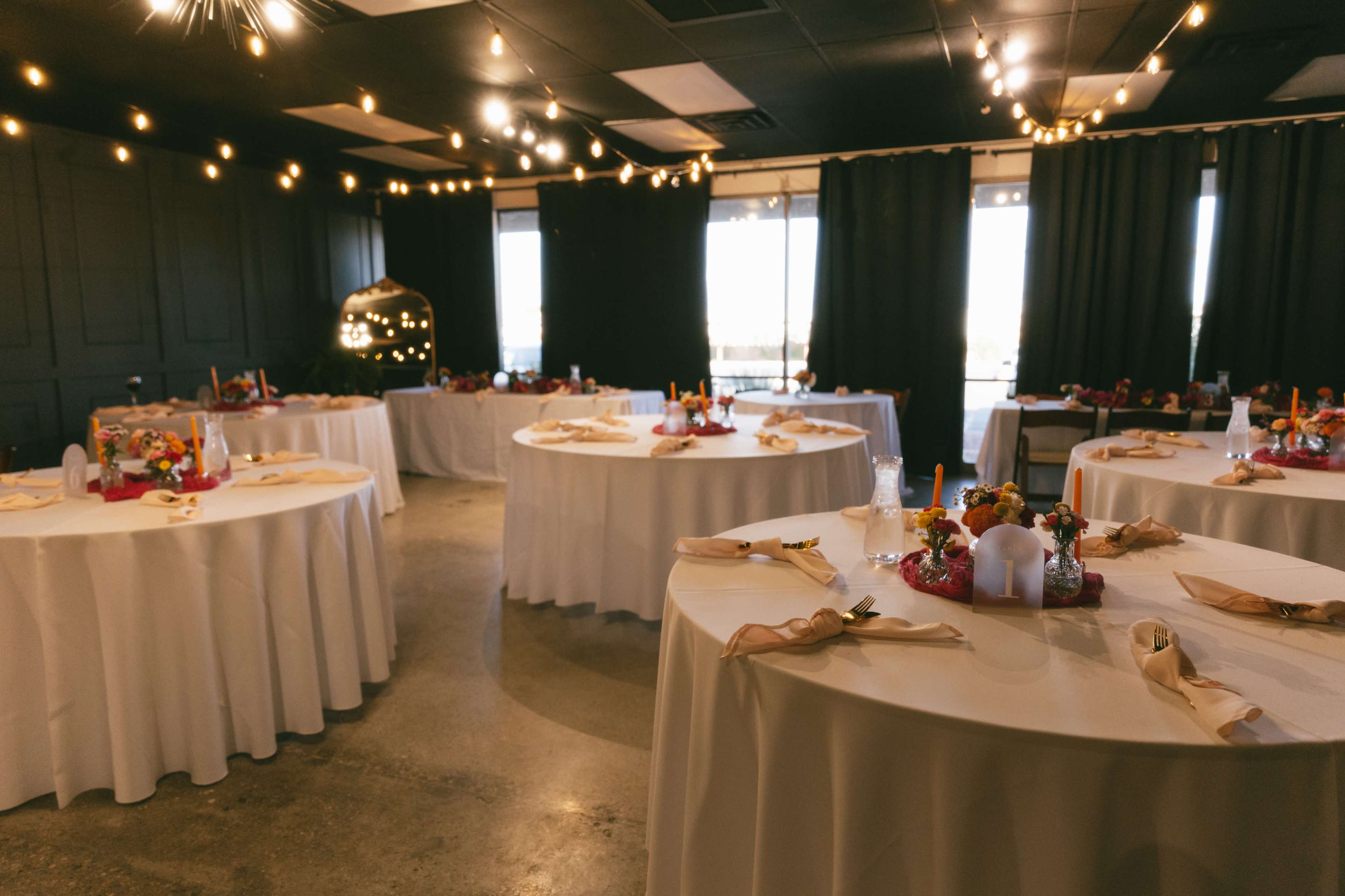 A banquet room with several circular tables set with white tablecloths and colorful centerpieces, illuminated by decorative string lights.